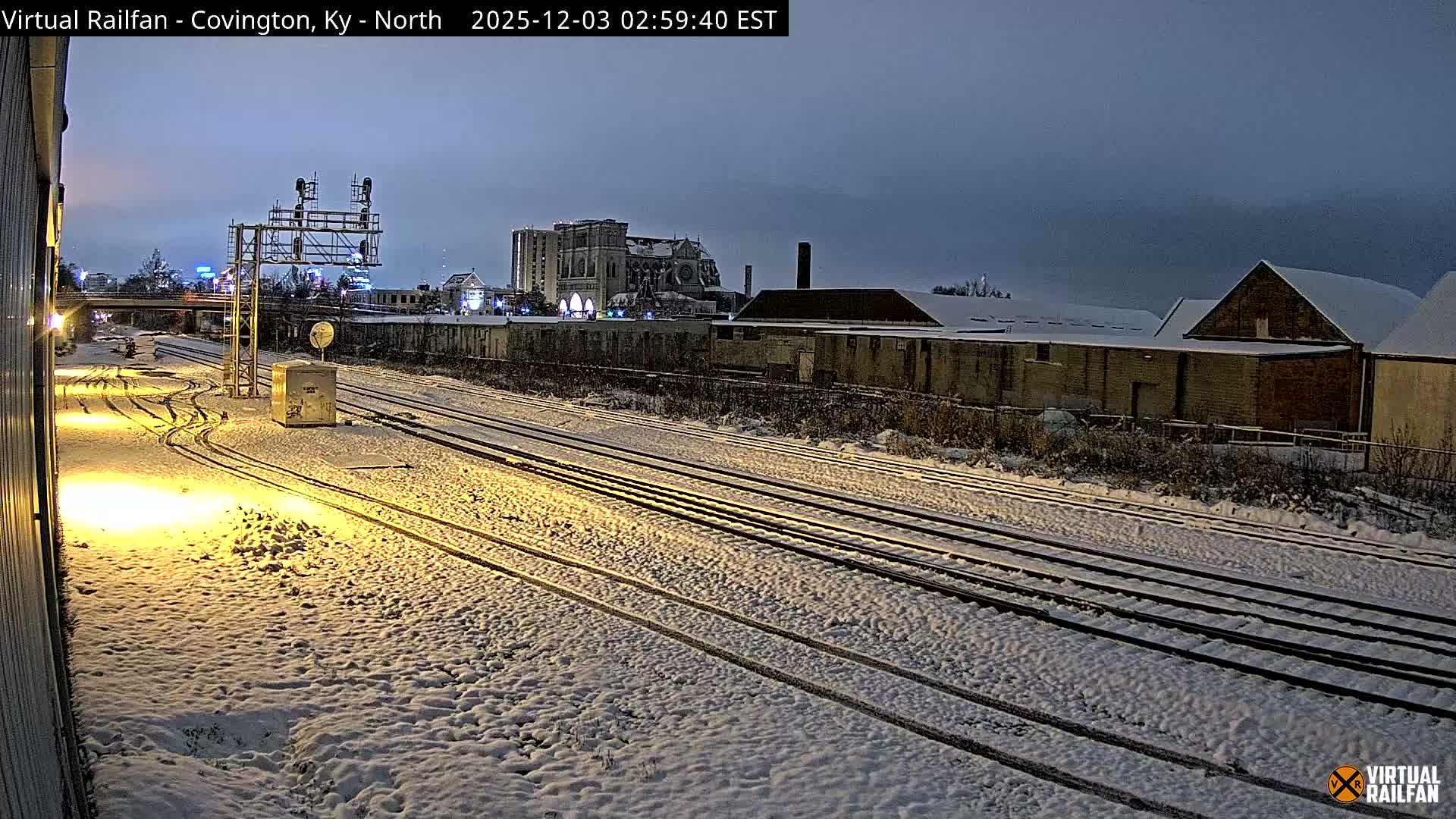 A nighttime or early morning view of a snow-covered railyard shows multiple train tracks illuminated by bright artificial lights, with industrial buildings and a dark, overcast sky overhead.