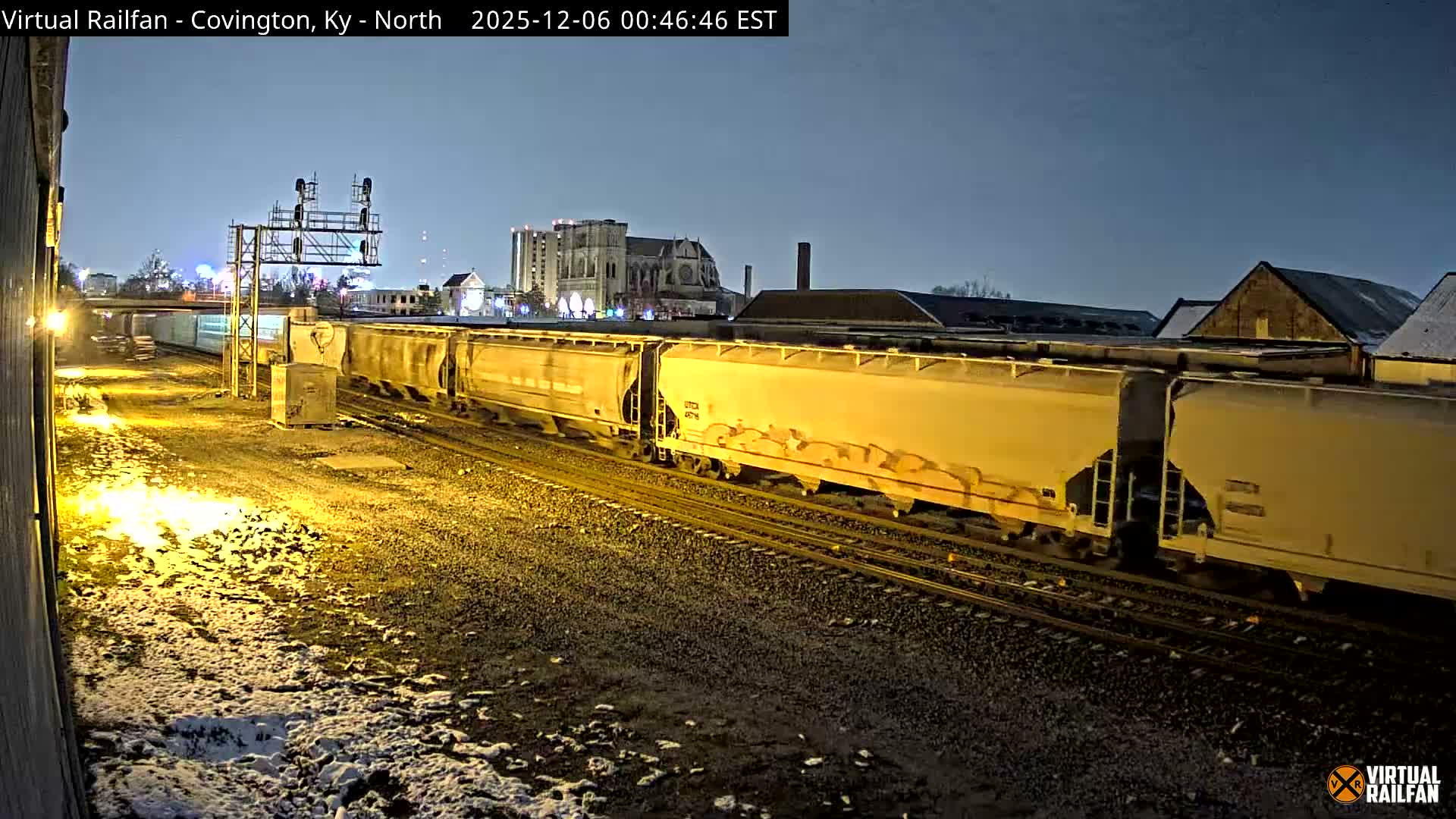 A long freight train with light-colored hopper cars is parked on snow-dusted tracks at night, brightly illuminated by artificial light, with a signal gantry and city buildings visible under a clear, dark sky.