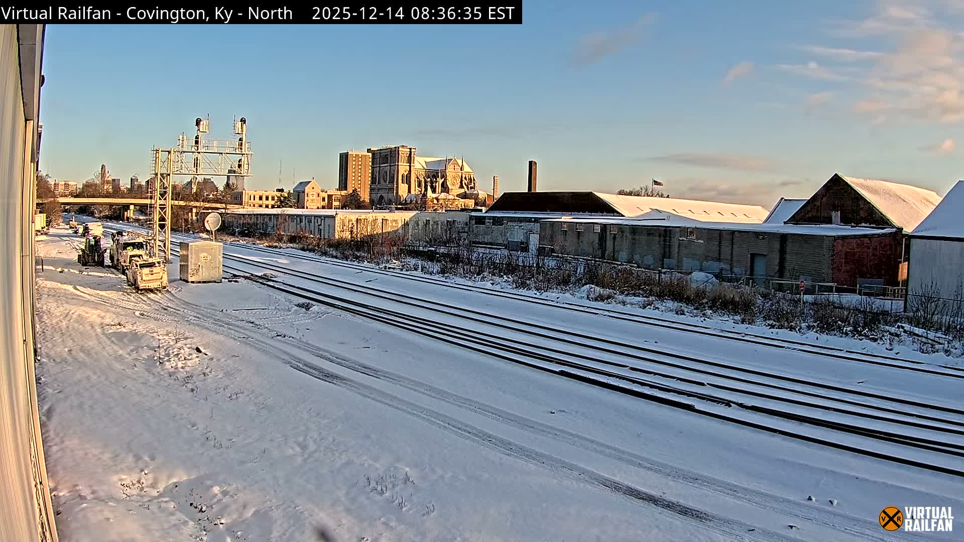 A long freight train with light-colored hopper cars is parked on snow-dusted tracks at night, brightly illuminated by artificial light, with a signal gantry and city buildings visible under a clear, dark sky.