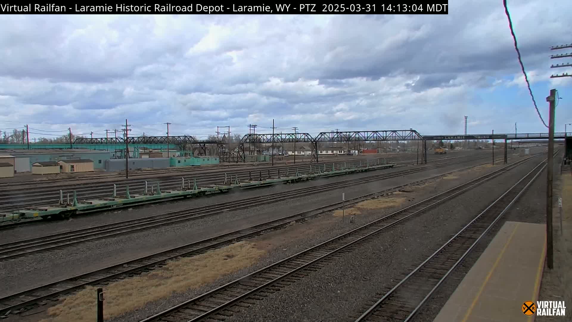 A wide shot of a railway yard under a cloudy sky shows numerous train cars and parallel tracks extending into the distance.