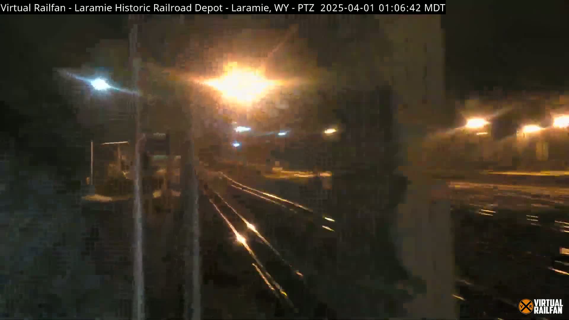 A nighttime view of railroad tracks at a depot, illuminated by streetlights under wet conditions.