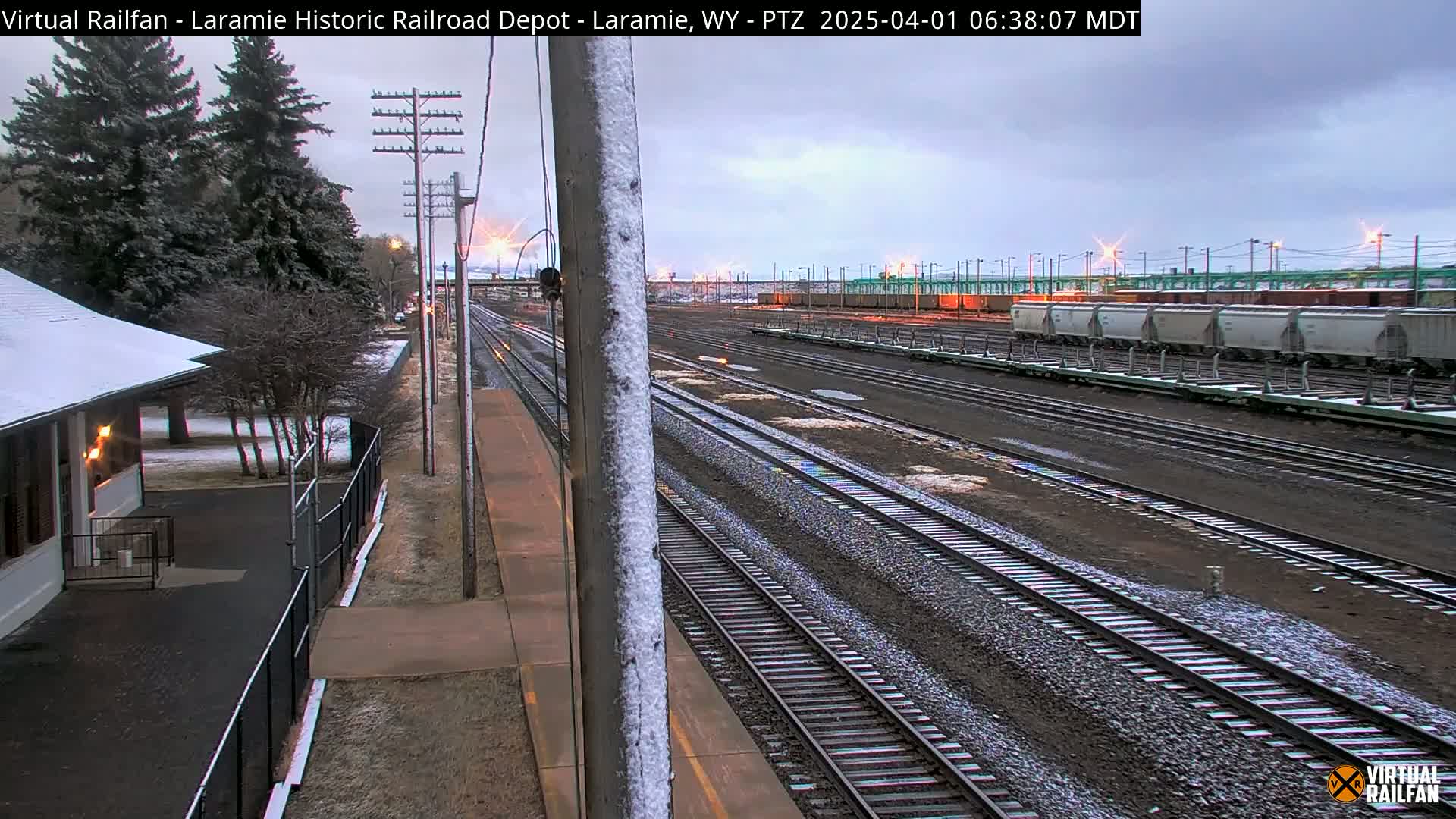 A snow-dusted train depot and numerous railway tracks extend into the distance under an overcast sky.