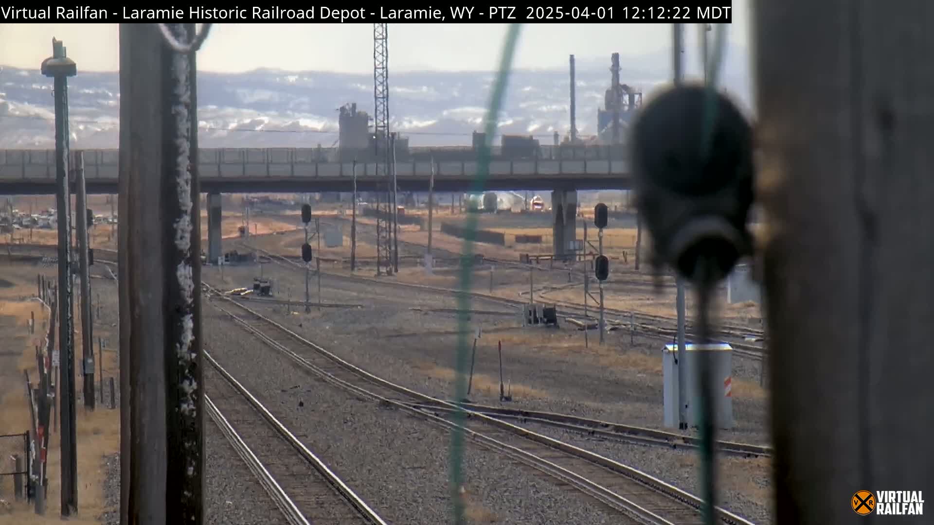 A view of multiple railroad tracks converging under a bridge on a hazy day, with an industrial area visible in the background.