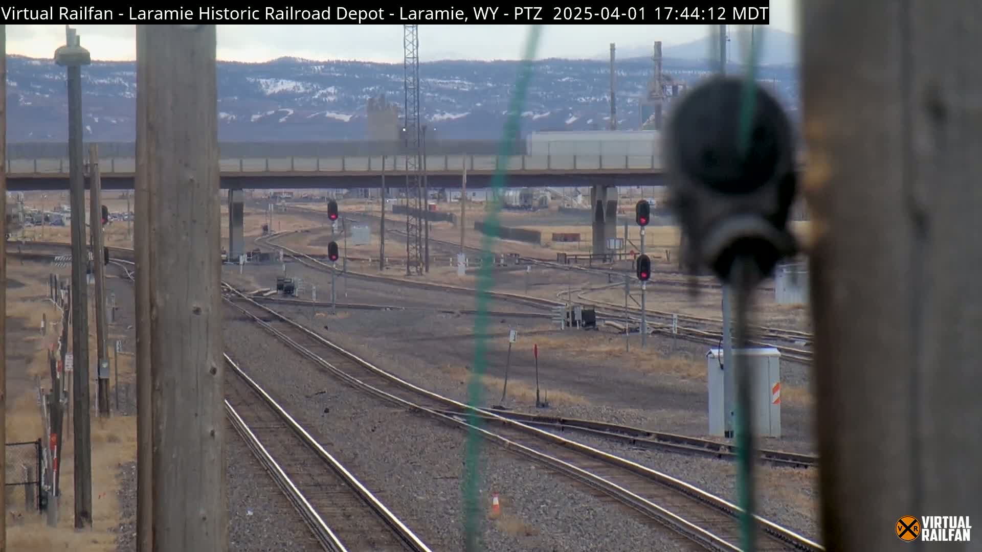 A view of multiple sets of train tracks intersecting under a bridge on a clear day, with distant mountains and industrial structures visible in the background.