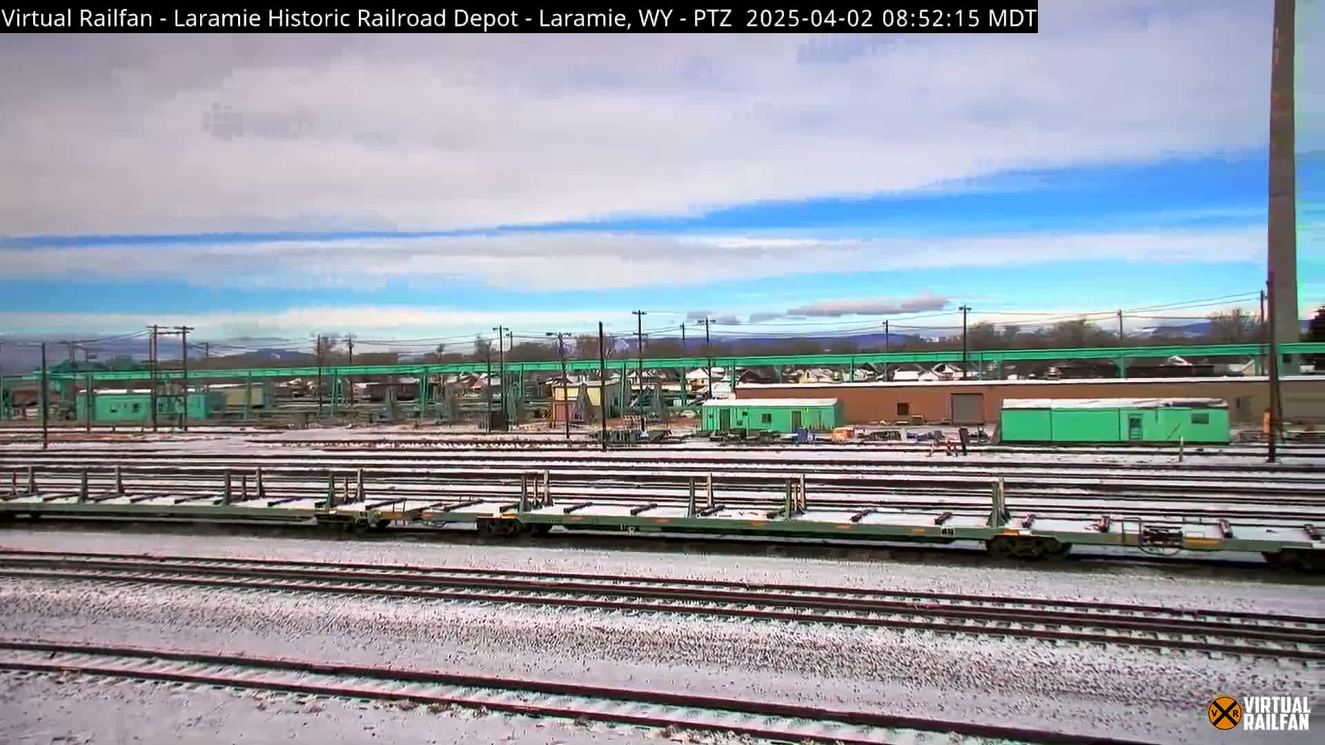 A snow-dusted rail yard with several flatbed train cars sits under a partly cloudy sky.