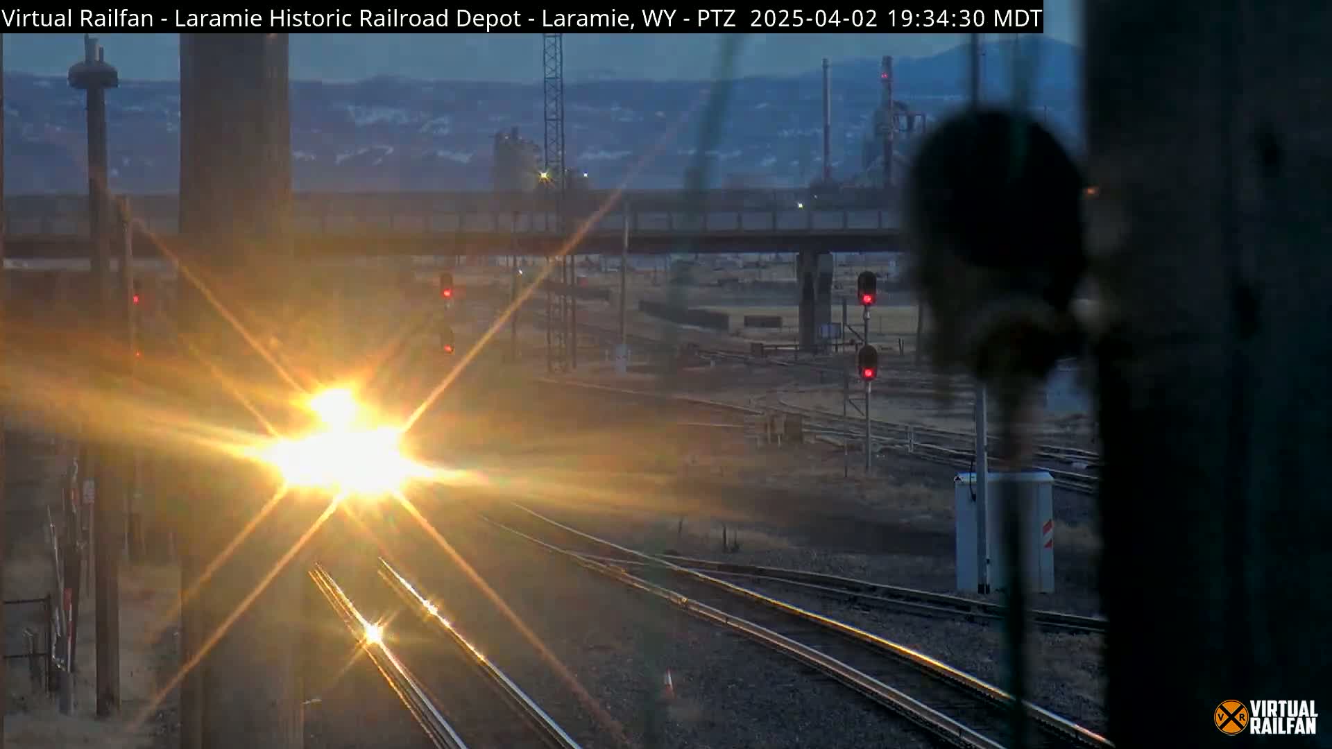 A bright, low-angled sun shines on a train approaching a railroad junction at dusk.