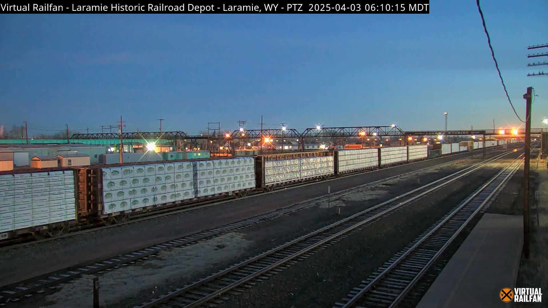 A long freight train carrying stacked packages is parked on multiple railroad tracks at twilight under a clear, dusky sky.