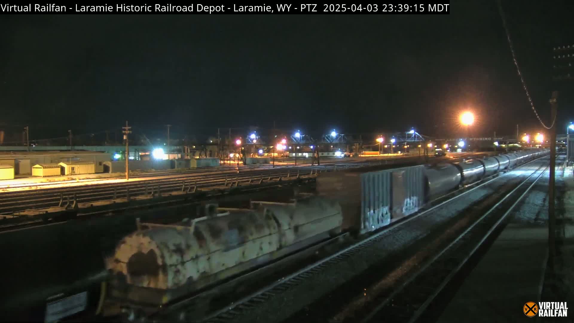 A long freight train moves through a rail yard at night in snowy conditions.