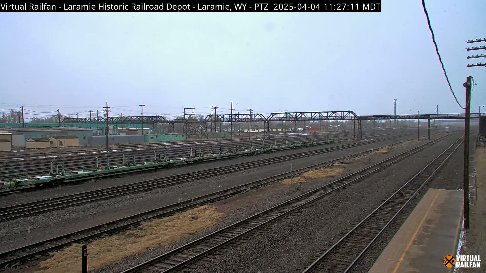 A long view of a railway yard with several parallel tracks, a train of light green freight cars, and a metal bridge under a lightly overcast sky.