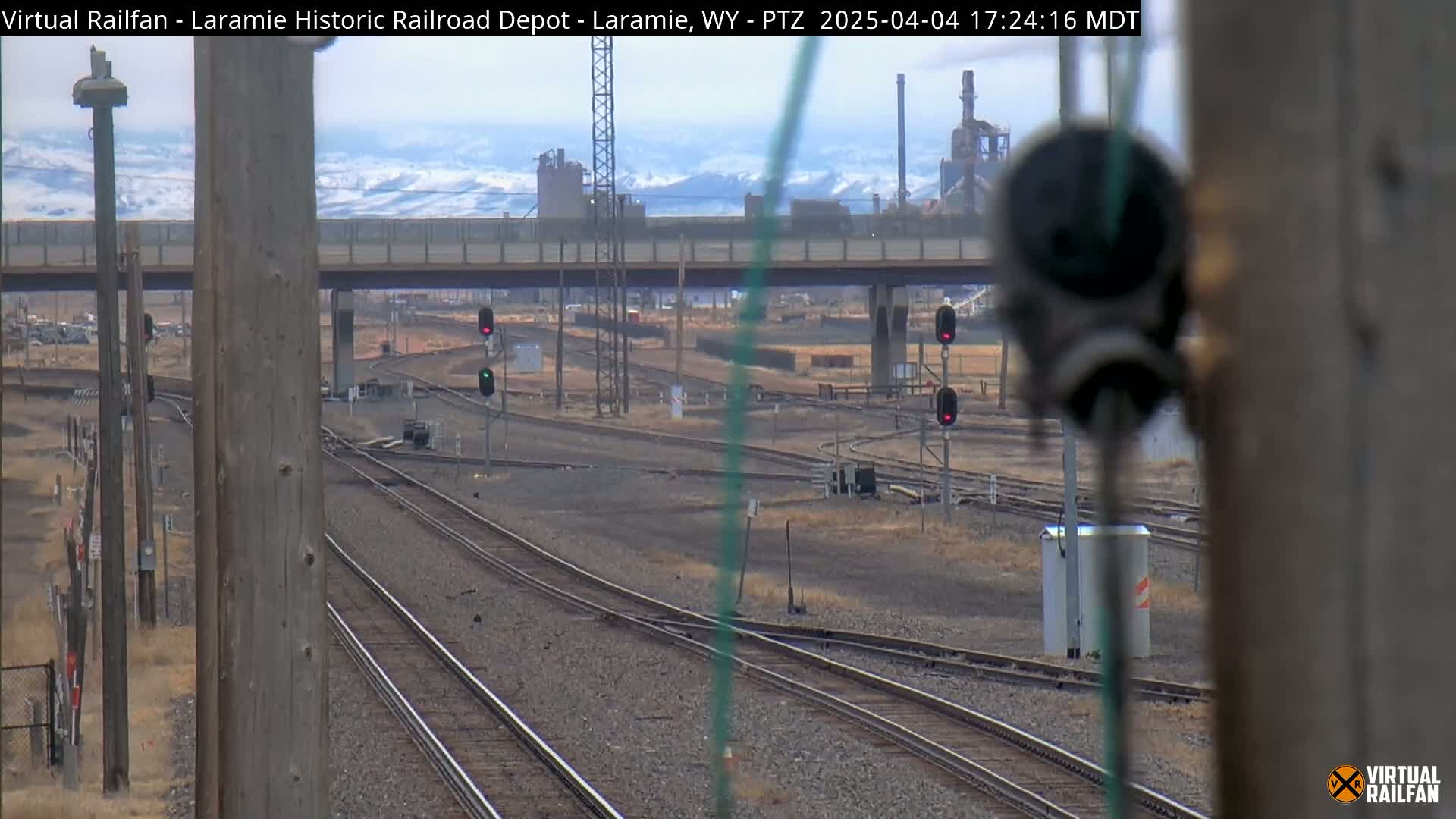 A view of railroad tracks and switching yards under a partly cloudy sky with snow-covered mountains in the distance.