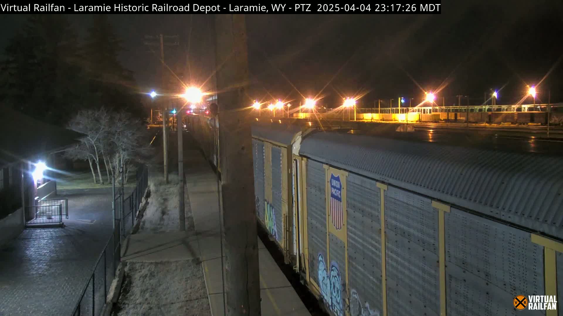 At night, a portion of a Union Pacific freight train sits alongside a paved walkway next to a dark, tree-lined area under illuminated streetlights.