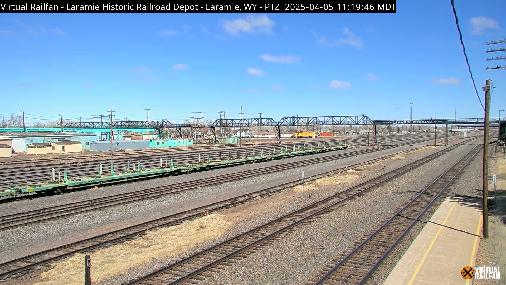 Under a clear blue sky, several light teal railroad cars sit on a vast network of train tracks next to industrial buildings.
