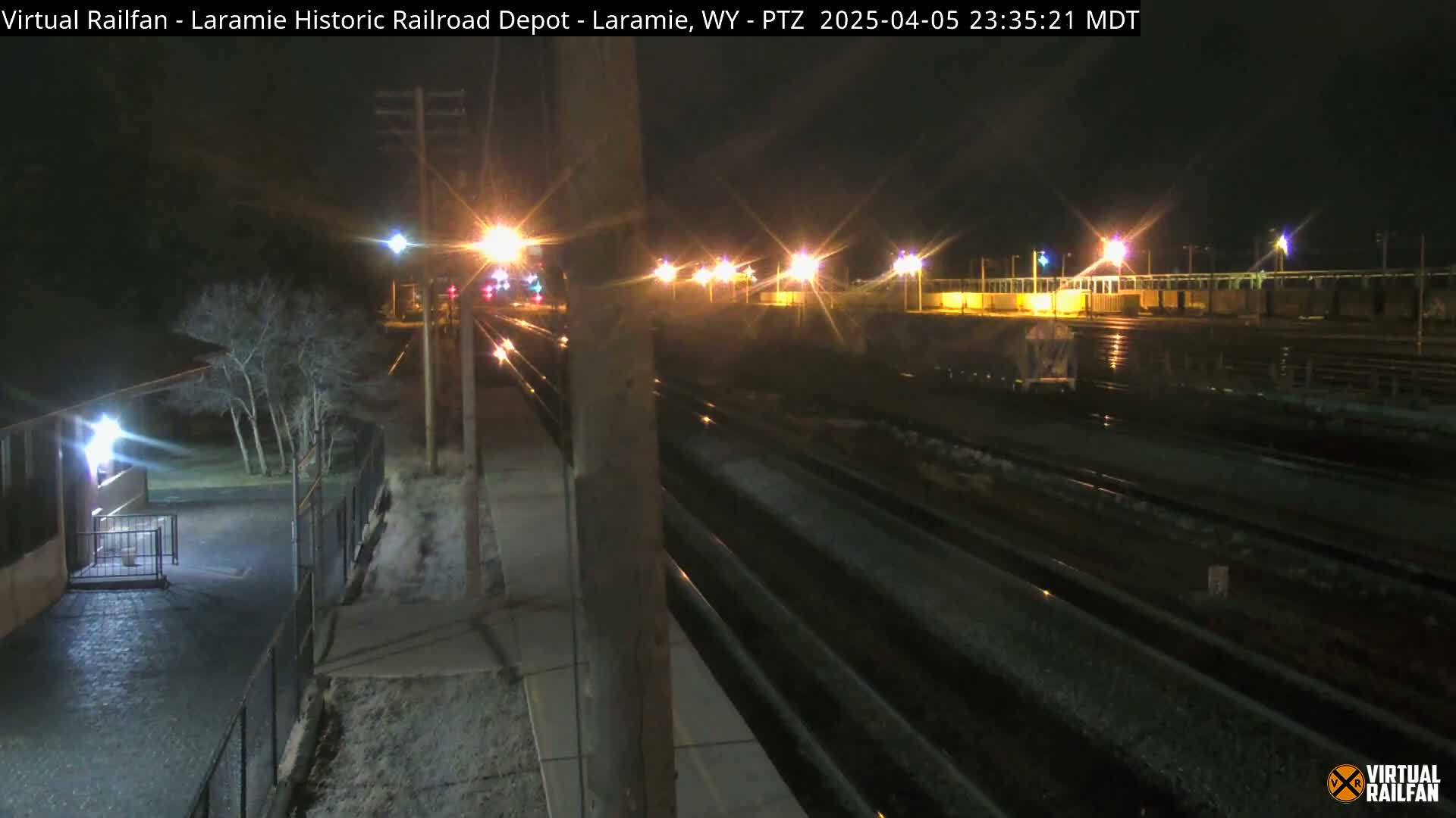 A nighttime view of railroad tracks and a street, illuminated by streetlights, with some light snow or ice visible on the ground.