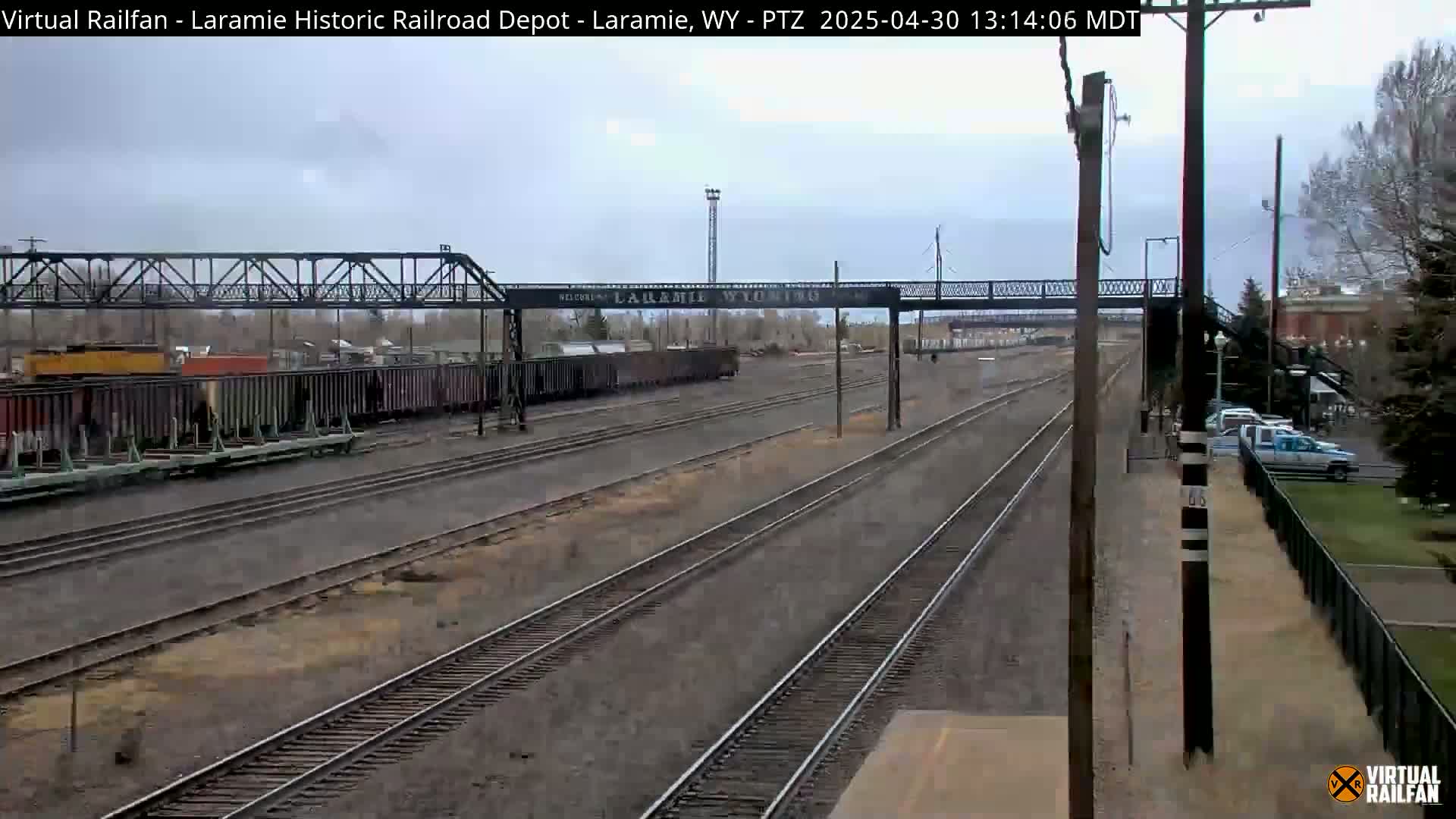 A railroad yard with multiple train tracks under an overcast sky, featuring a metal pedestrian bridge and a line of freight cars.