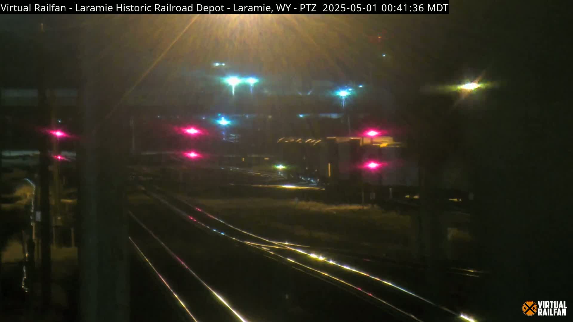 At night, a train passes by several railroad tracks under hazy conditions, with lights illuminating the scene.