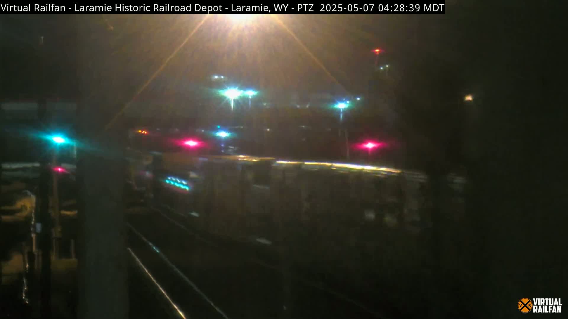 A nighttime view of a long, dark train moving past illuminated lights and signals.