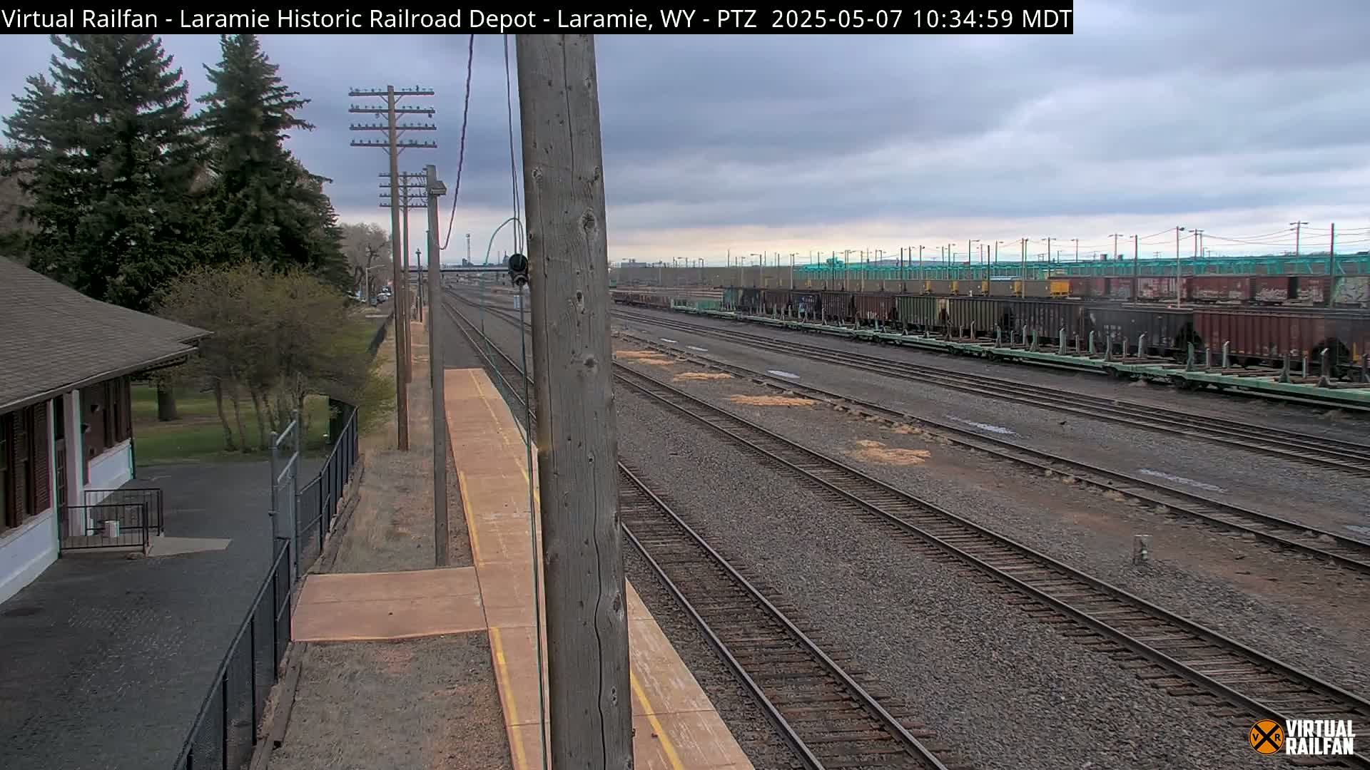 A long line of freight cars sits on a railway under a cloudy sky, next to a small depot and trees.