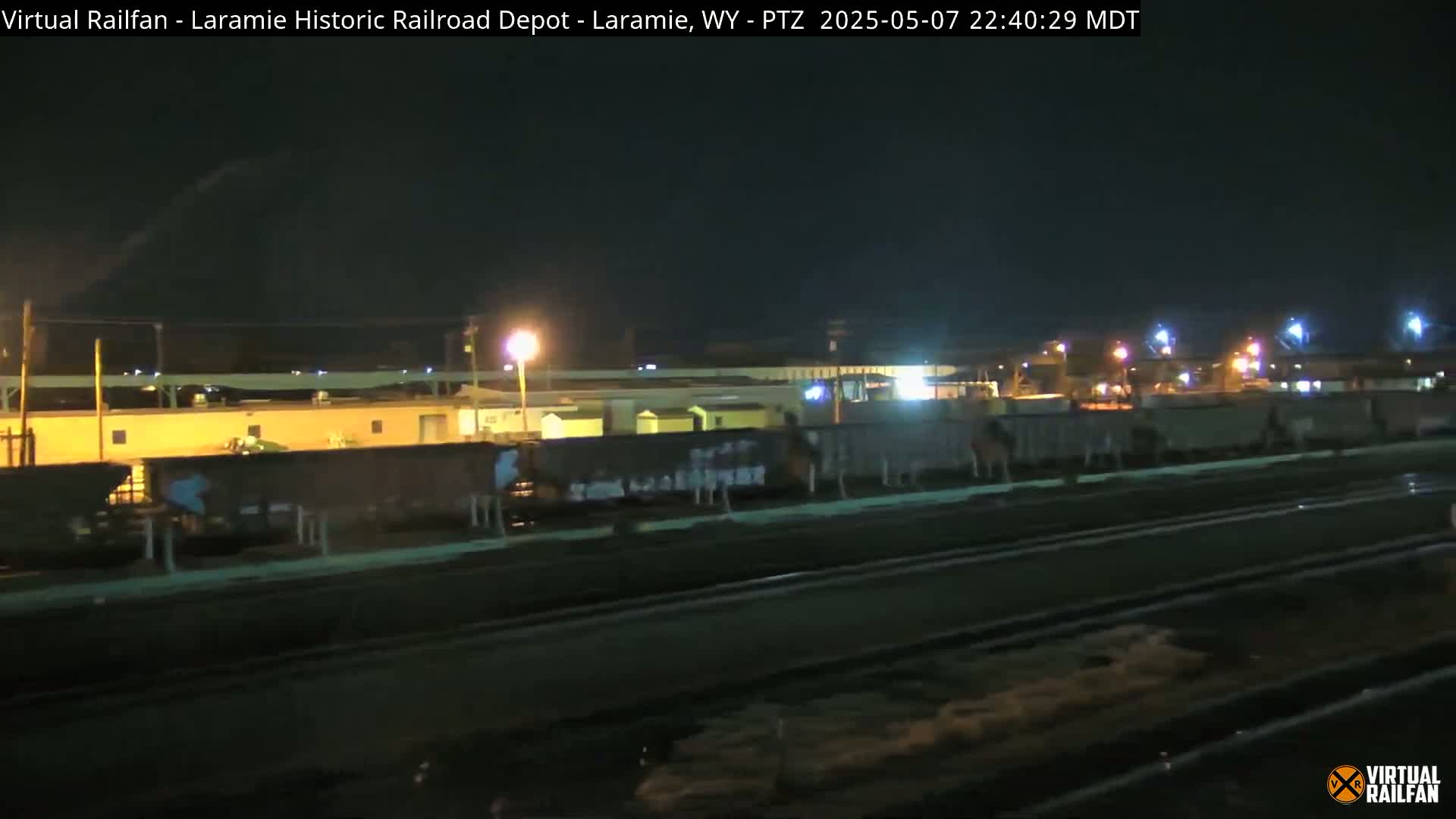 At night, a long freight train sits stationary on railroad tracks, under a dark sky, in front of a brightly lit building.