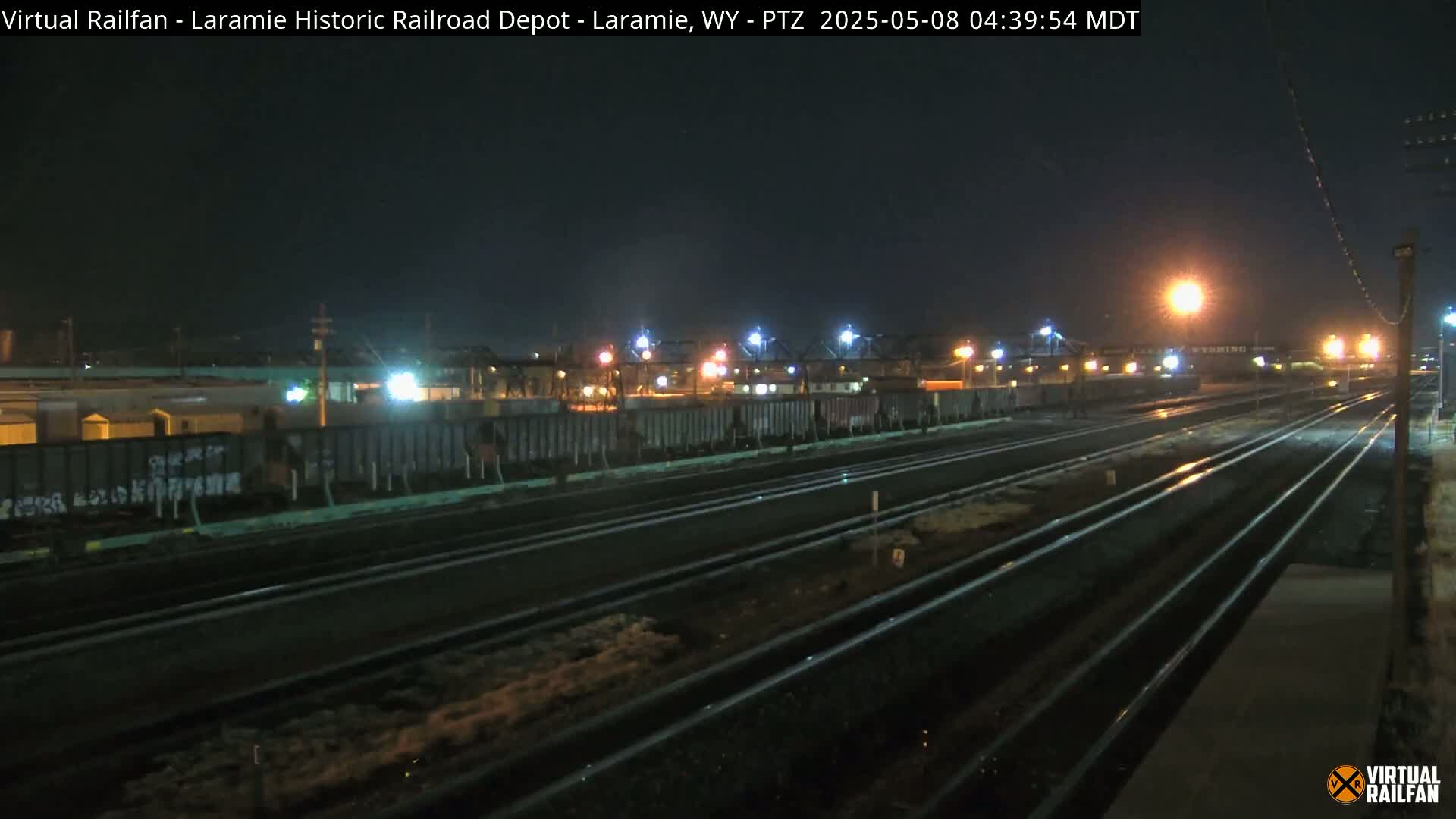 At night, a railroad yard with multiple sets of tracks and a line of boxcars is illuminated by artificial lights.