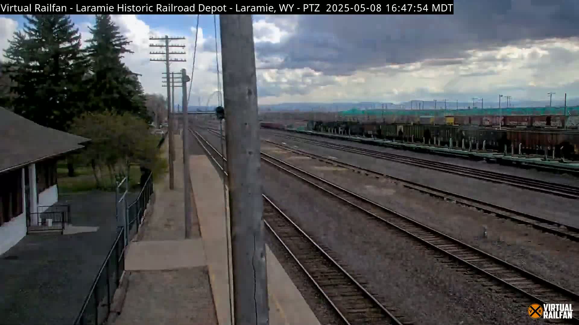 A railroad depot and several parallel train tracks extend into the distance under a cloudy sky.