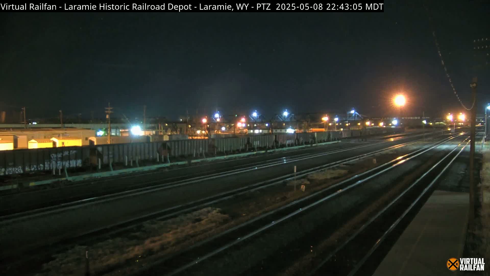 A nighttime view of a railroad yard with several train cars and multiple sets of tracks, illuminated by artificial lights under a dark sky.