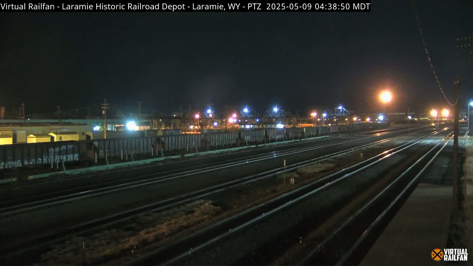 At night, a long line of dark rail cars sits beside multiple sets of train tracks under artificial lights.