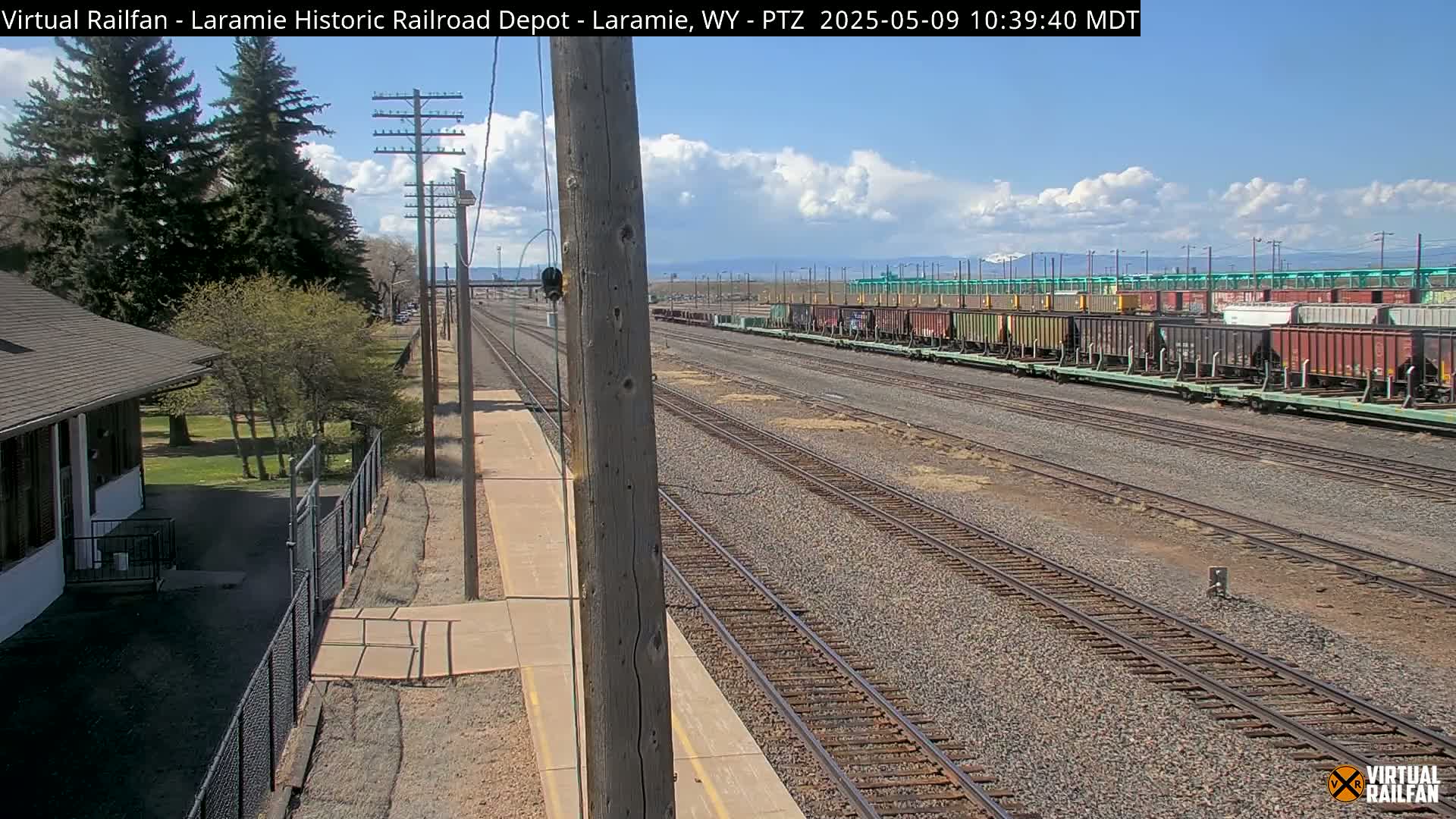 A long line of freight cars sits on railroad tracks under a partly cloudy sky, adjacent to a small depot building and several trees.