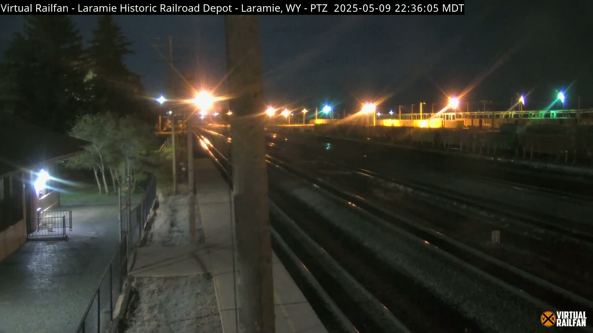At night, a wet railroad yard with multiple tracks is illuminated by streetlights, bordered by trees and a building.