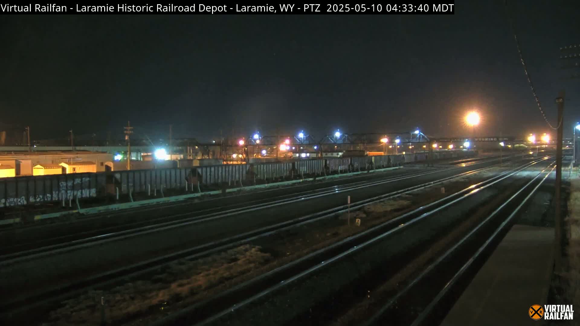 At night, a train yard with multiple sets of tracks shows several rail cars, some with graffiti, under artificial lighting.