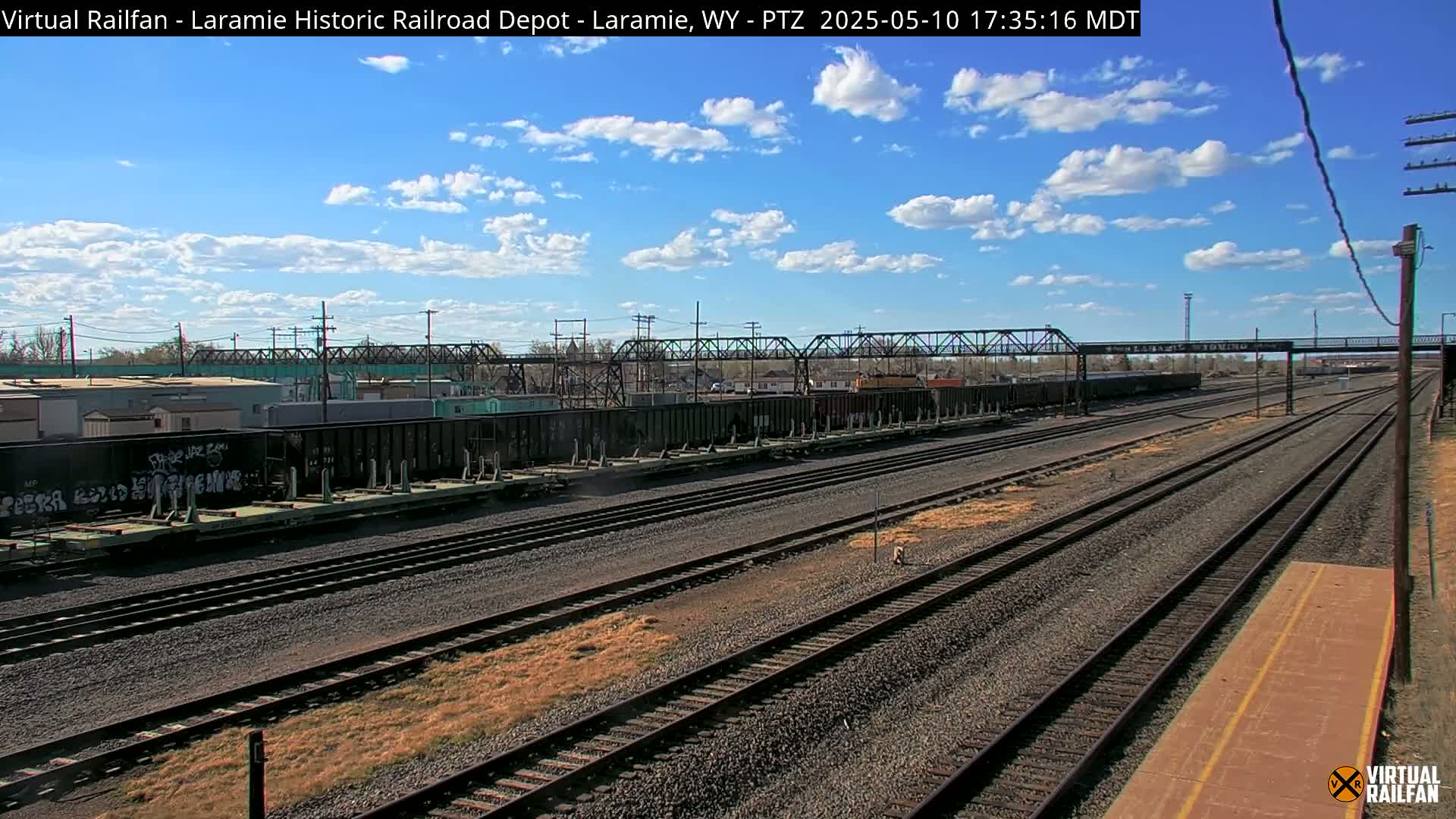 A sunny day with some clouds reveals a railroad yard with multiple train cars and tracks extending into the distance, under a metal bridge.