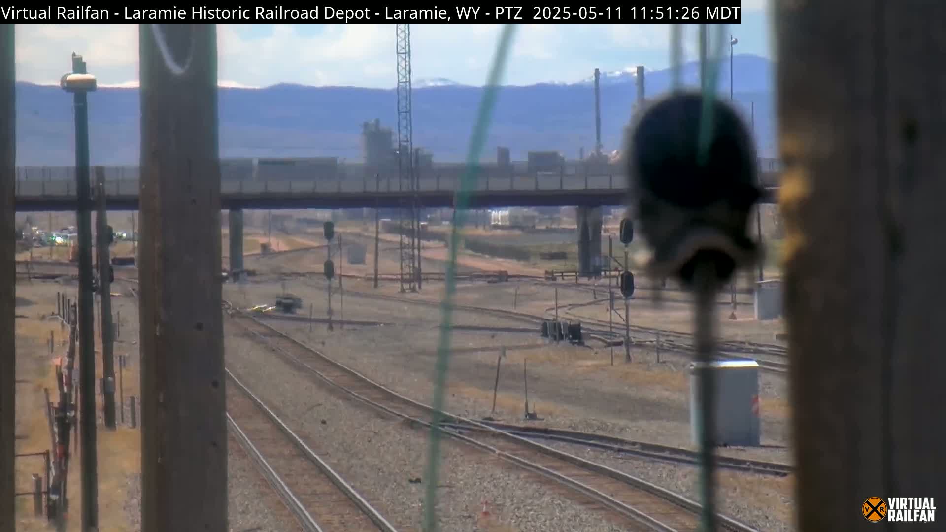A view of multiple train tracks, a highway overpass, and an industrial area in the distance under a clear sky.