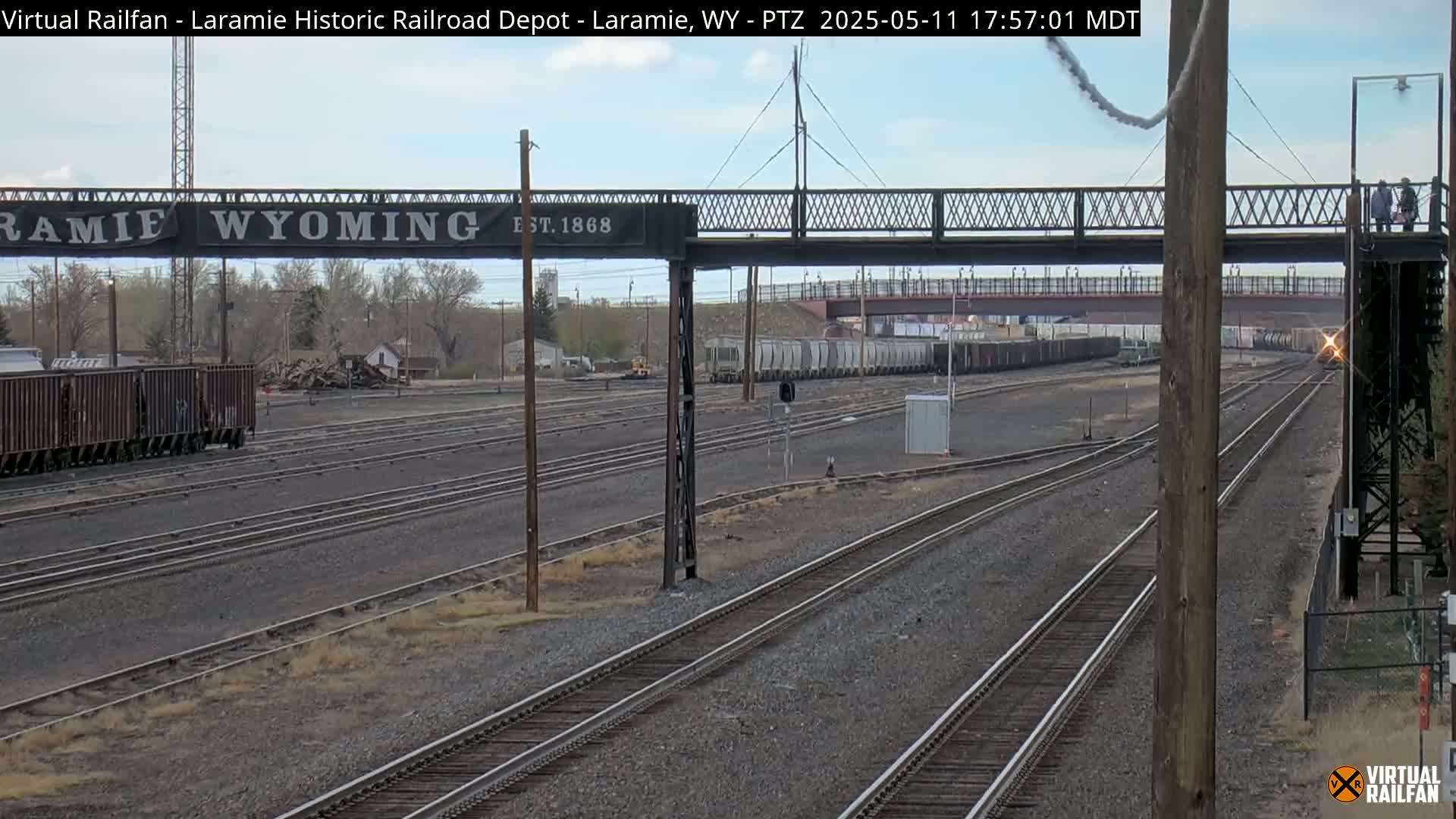 A train passes under a pedestrian bridge at a railroad yard on a partly cloudy day.