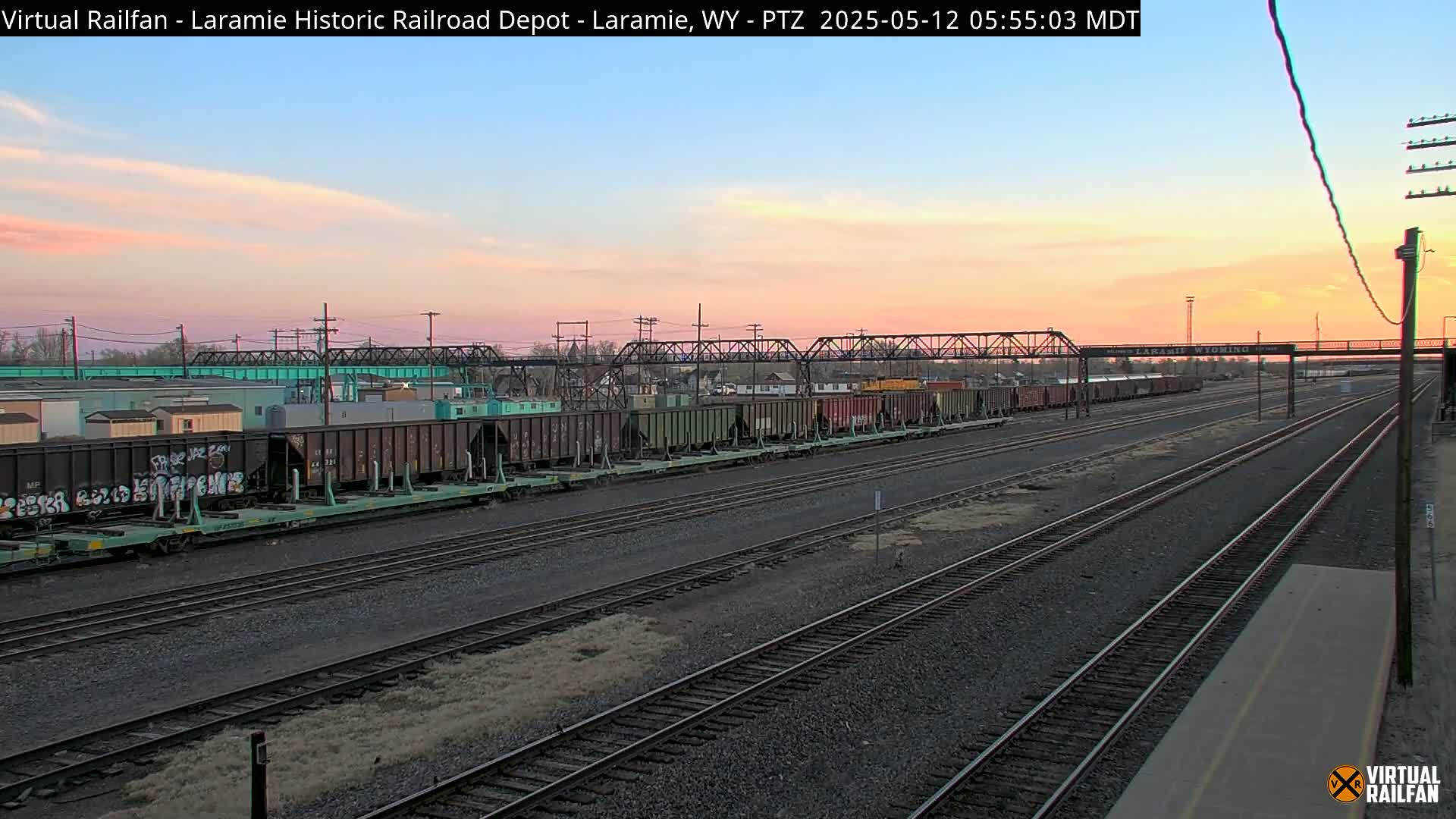 A long freight train sits on multiple parallel railway tracks at sunset under a pale pink and orange sky.