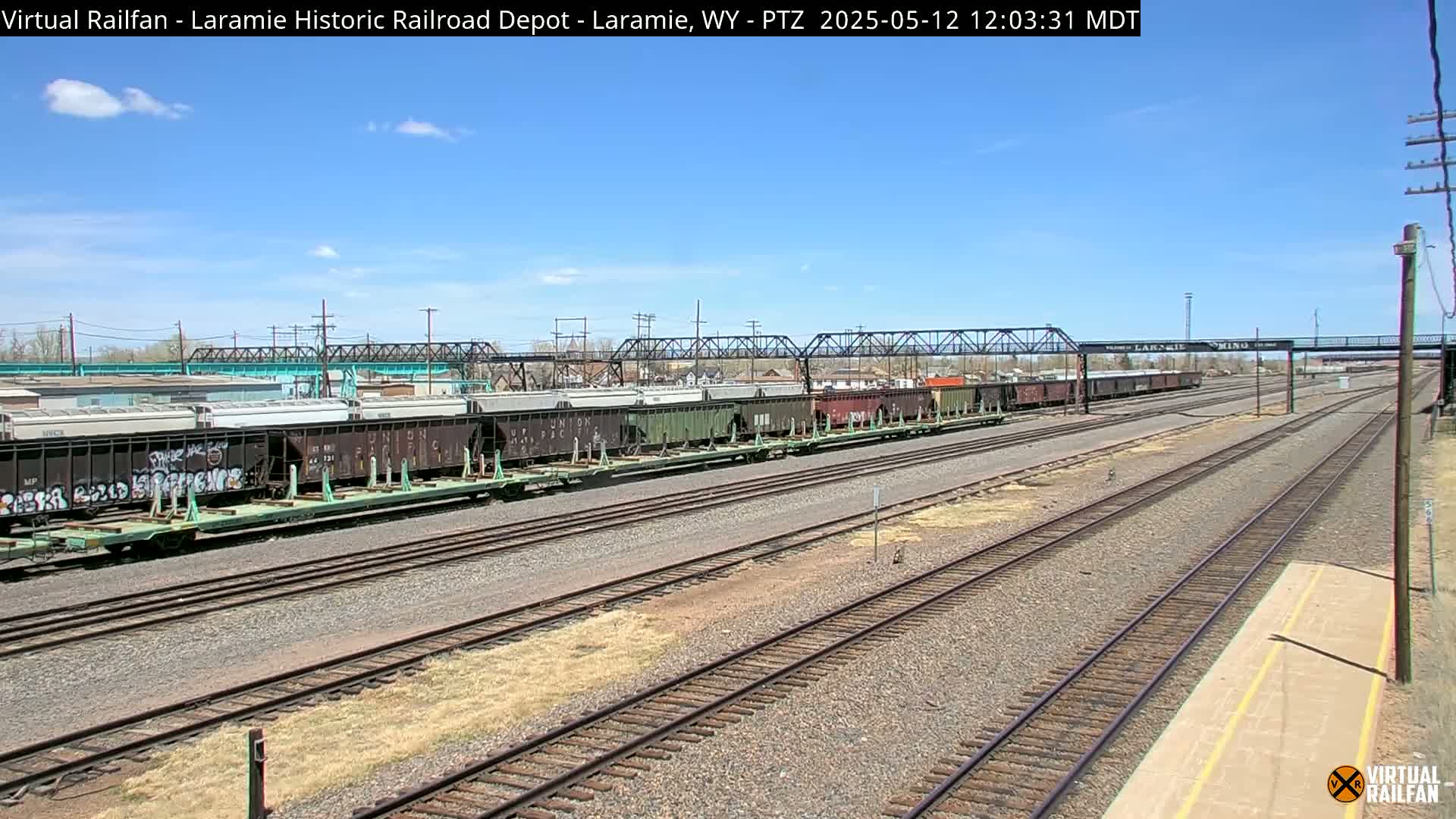 Under a clear blue sky, a long freight train sits on a set of railroad tracks, with additional tracks extending into the distance.
