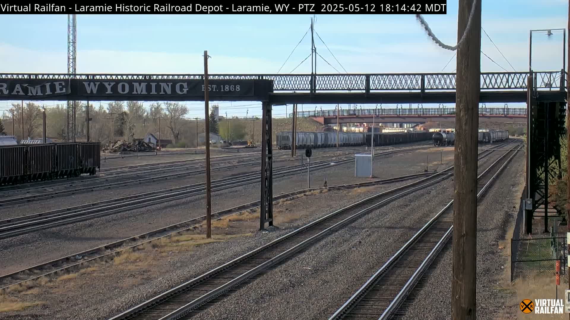 A train yard with multiple parallel train tracks, a railway bridge overhead, and a long freight train in the distance under a partly cloudy sky.