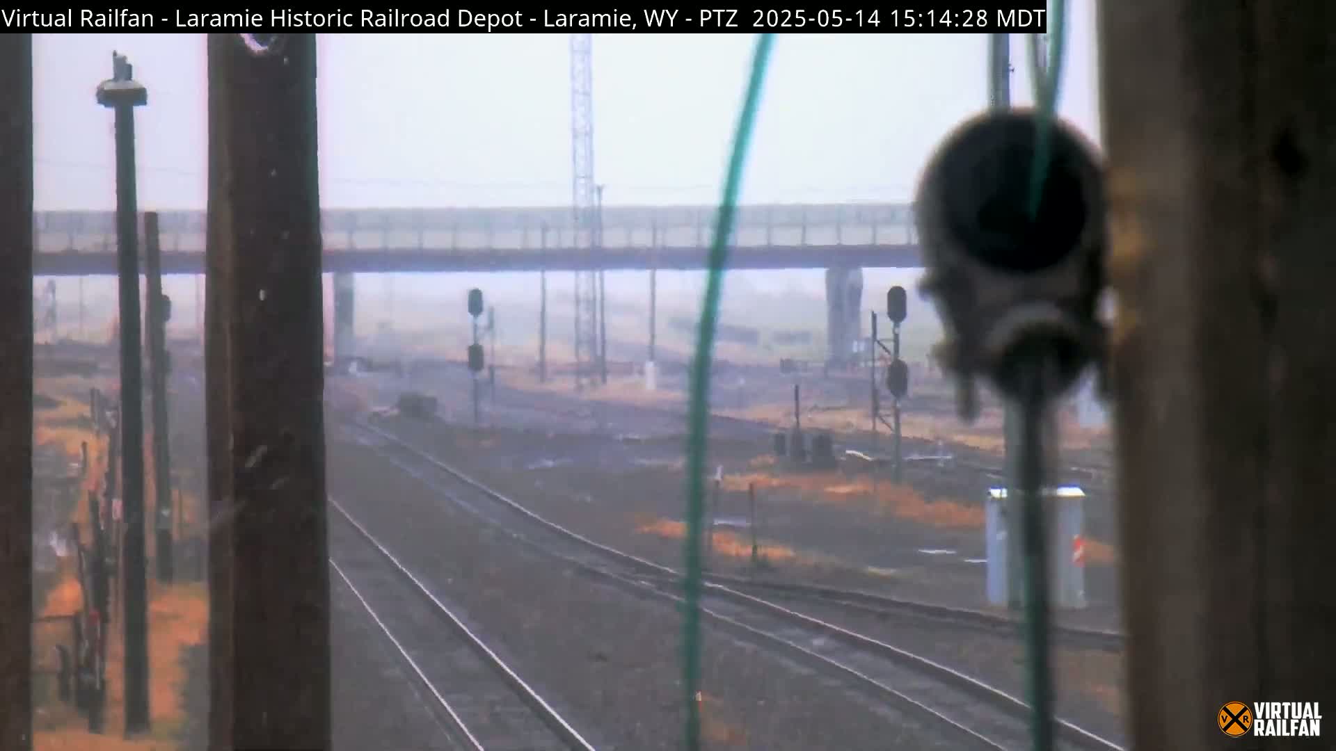 A view through a partially obscured window shows several train tracks extending into a hazy, overcast distance beneath a bridge.
