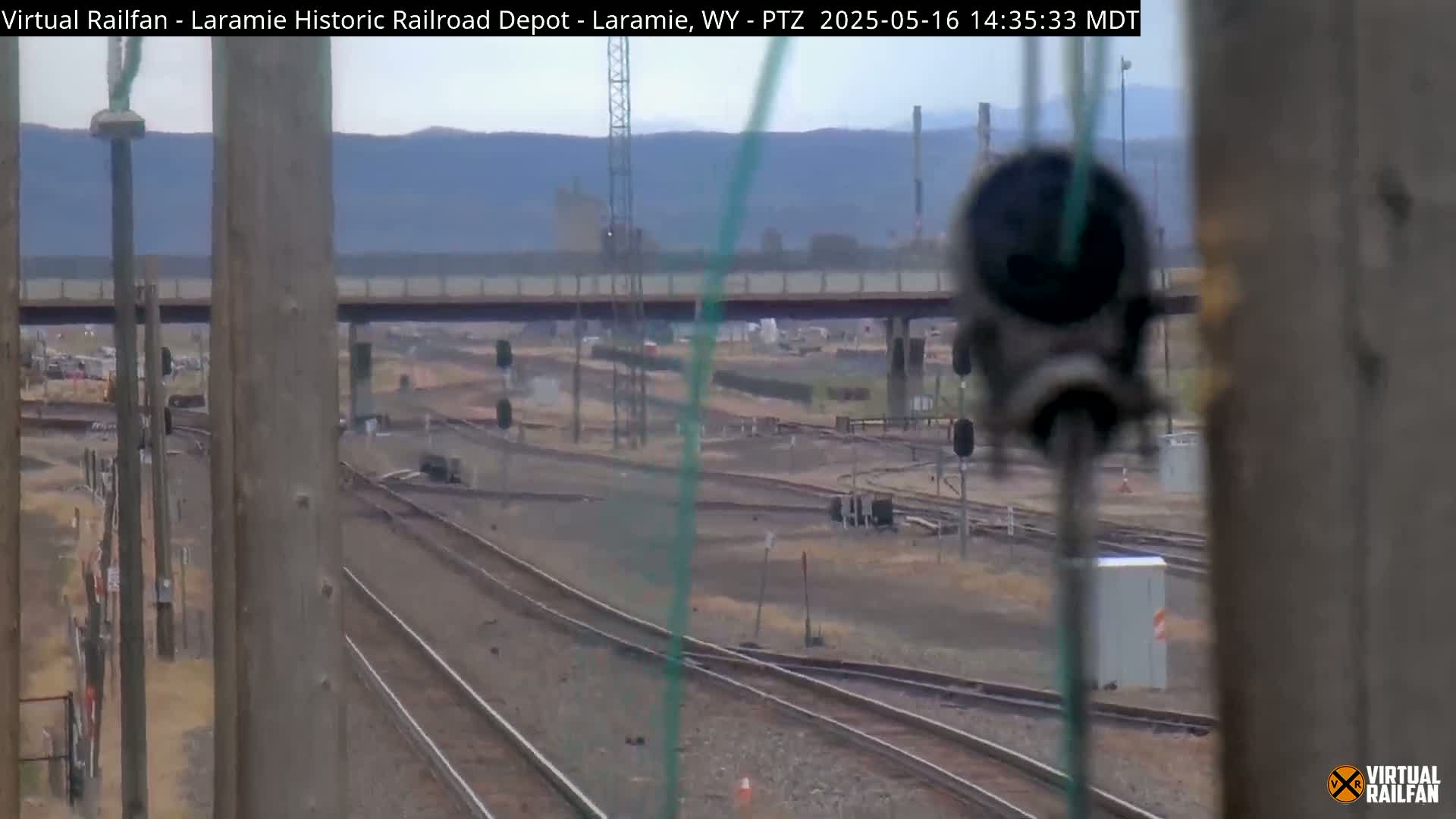 A view through wooden posts shows several railway tracks converging and diverging under an overcast sky, with a road overpass in the background.
