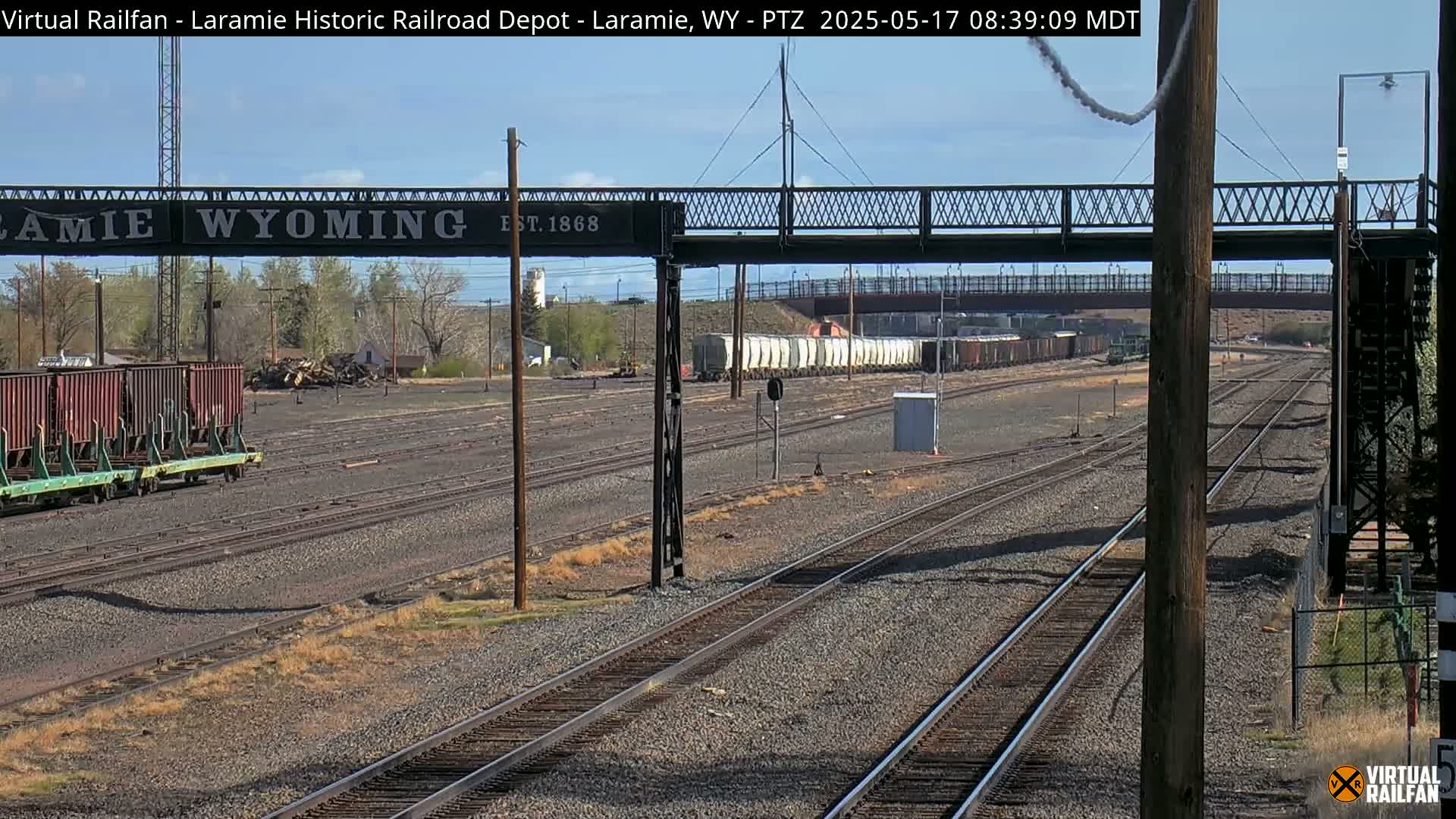 A railway yard with multiple train cars under a partly sunny sky, featuring a pedestrian bridge with a sign that reads “Laramie Wyoming.”