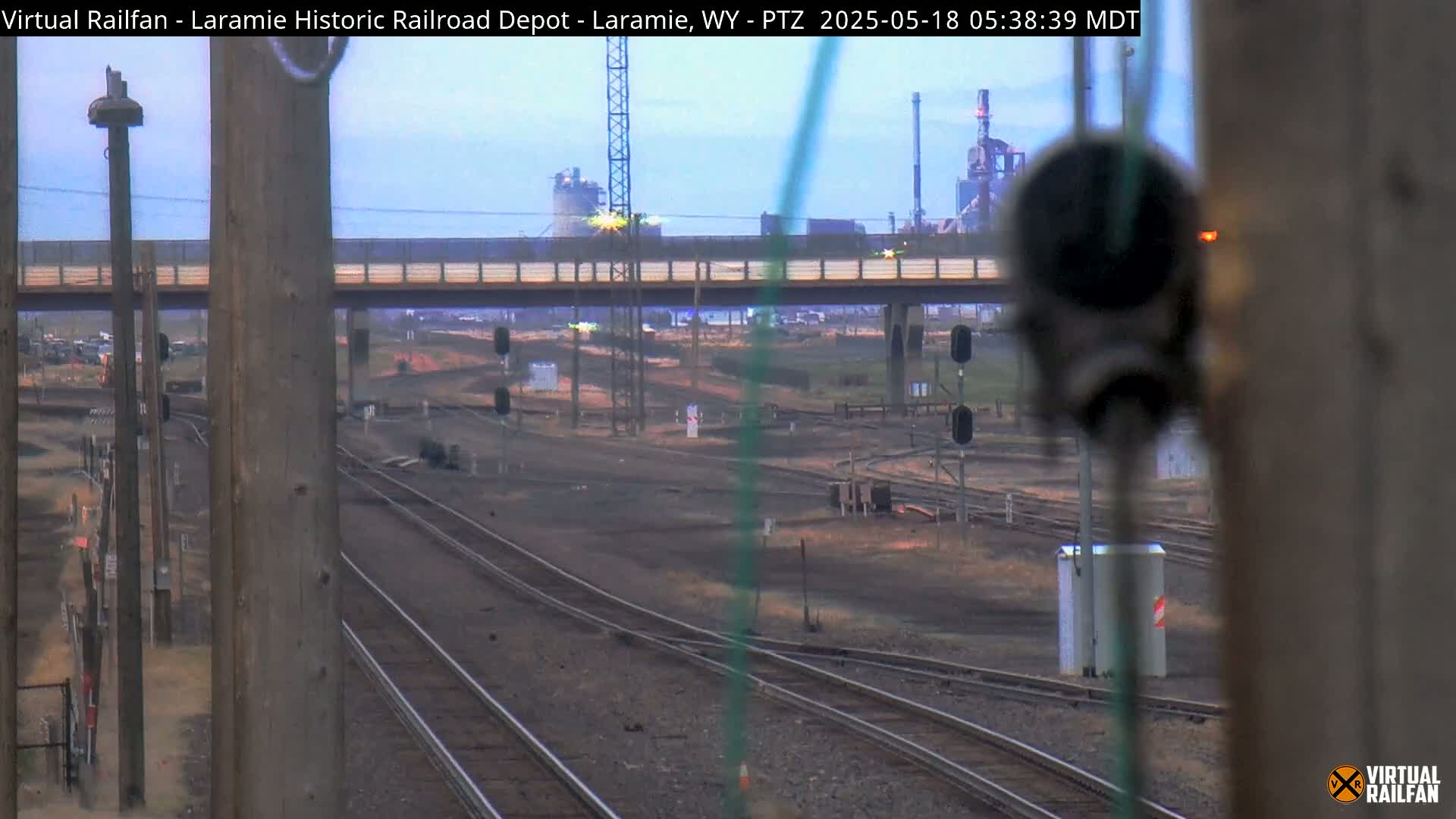Several sets of train tracks converge under a bridge and pass in front of an industrial plant under a mostly clear sky.