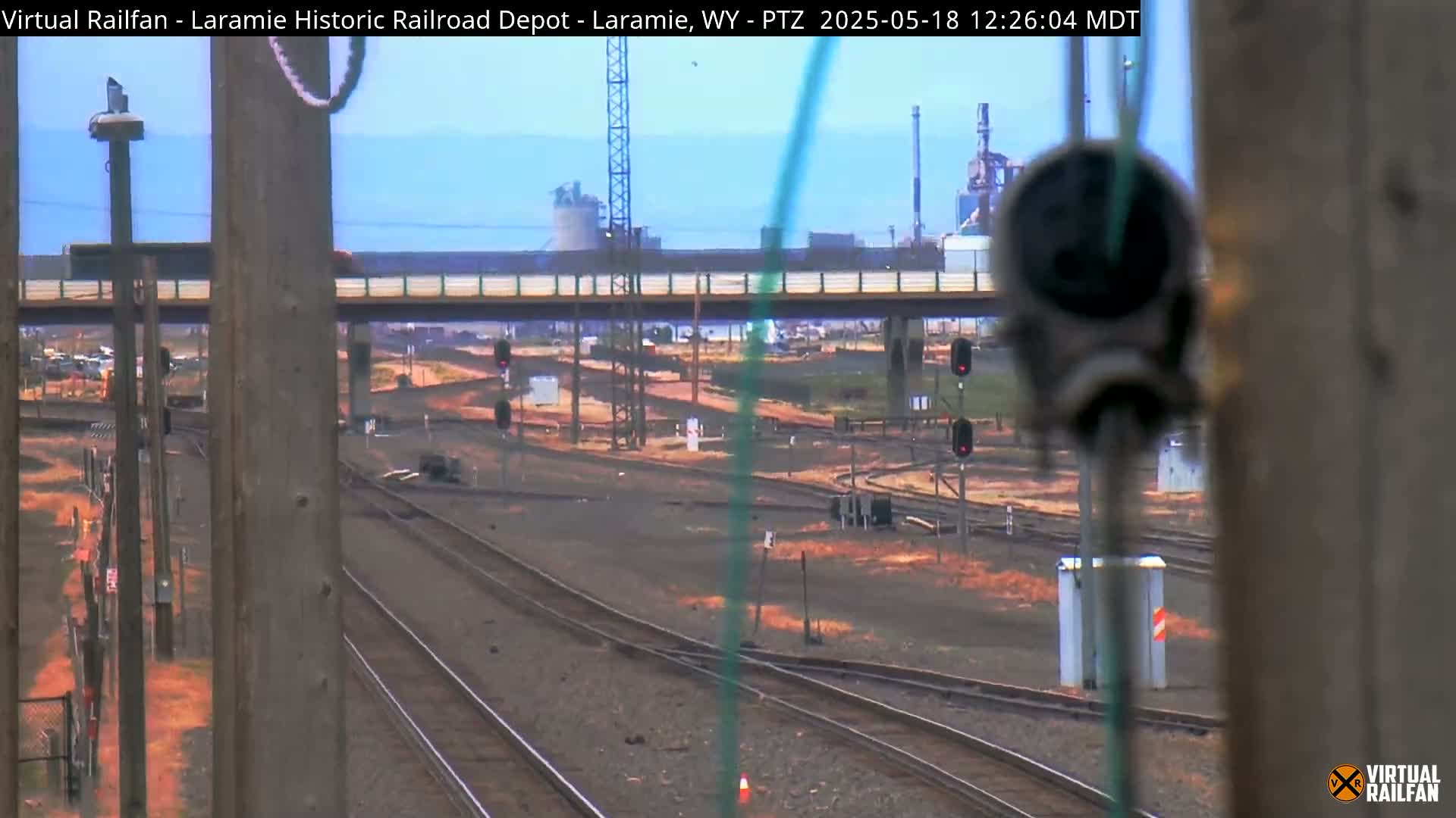 A view of railroad tracks and switching areas, with an industrial plant in the background on a partly cloudy day.