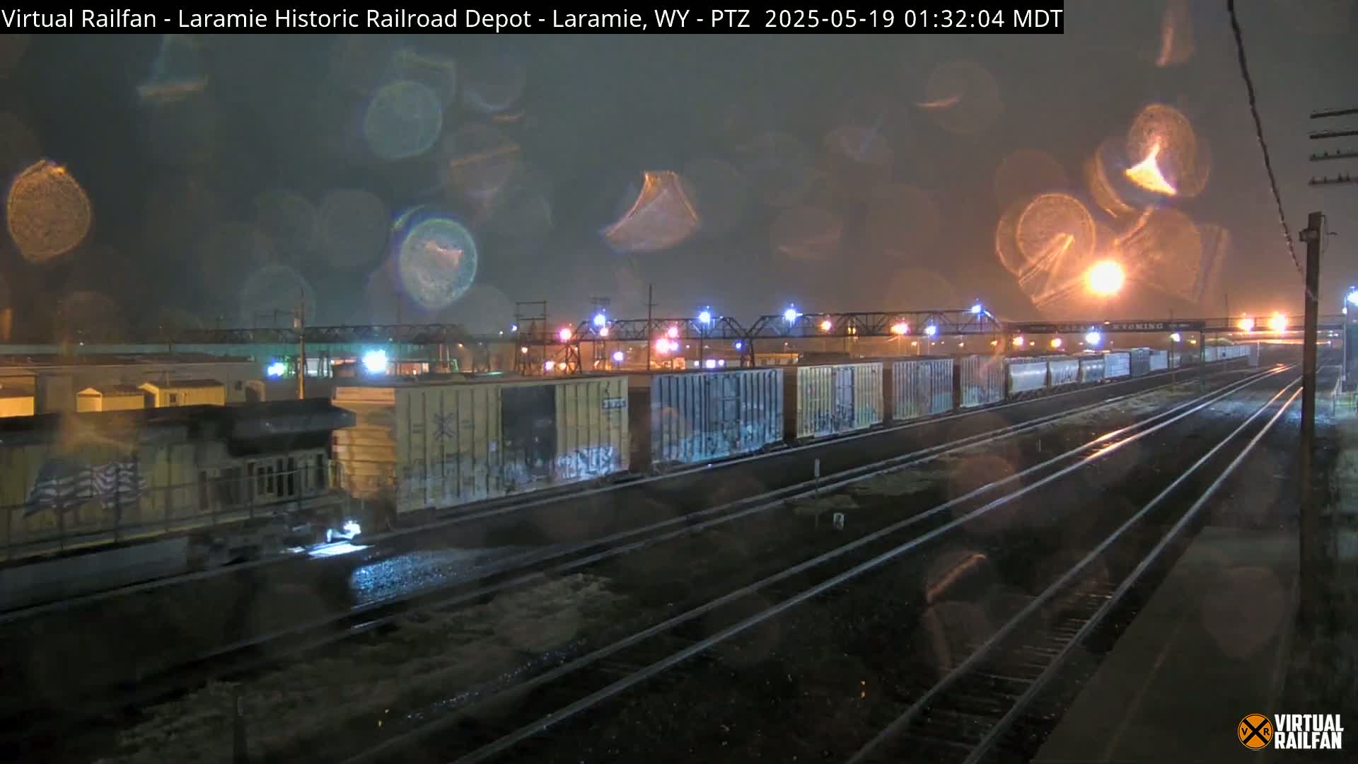A nighttime view of a train with graffiti-covered boxcars moving through a railyard during a rainstorm.