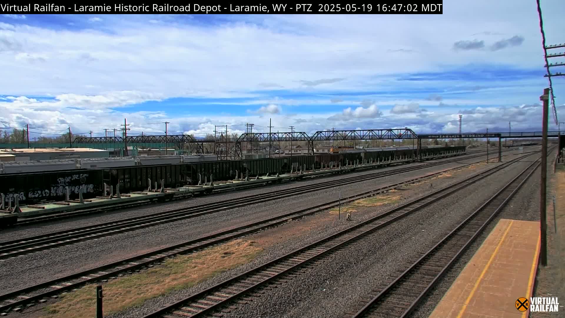 A mostly sunny day shows a long line of dark-colored freight cars parked on train tracks, with additional tracks extending into the distance under a partly cloudy sky.