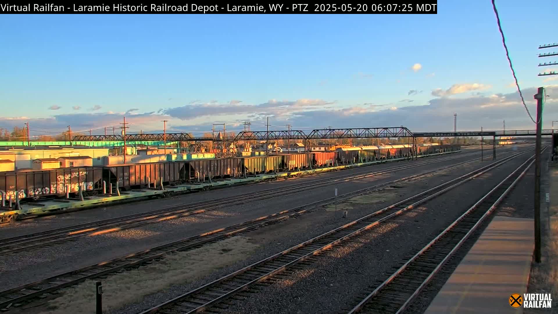 Under a mostly clear, late afternoon sky, a long line of freight train cars sits on a rail yard with multiple sets of tracks extending into the distance.