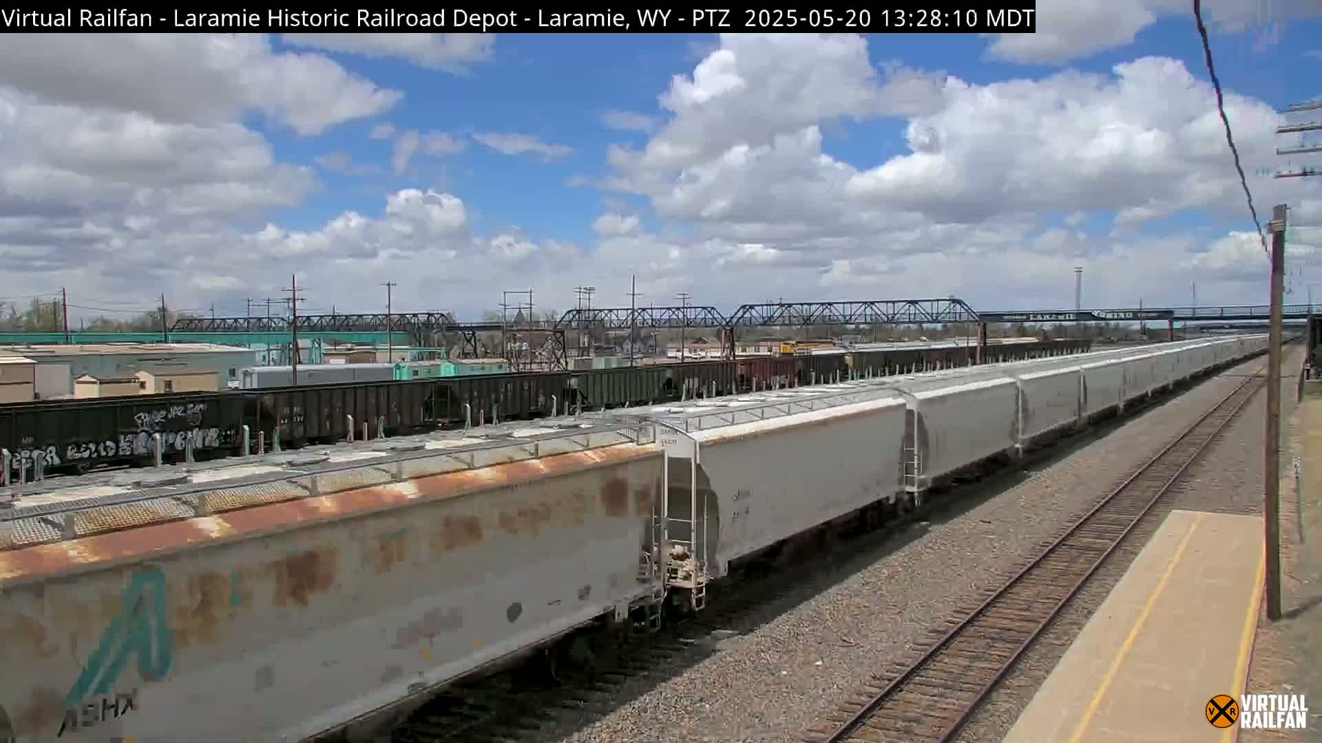 A long freight train of light gray boxcars sits on a railway track under a partly cloudy blue sky.