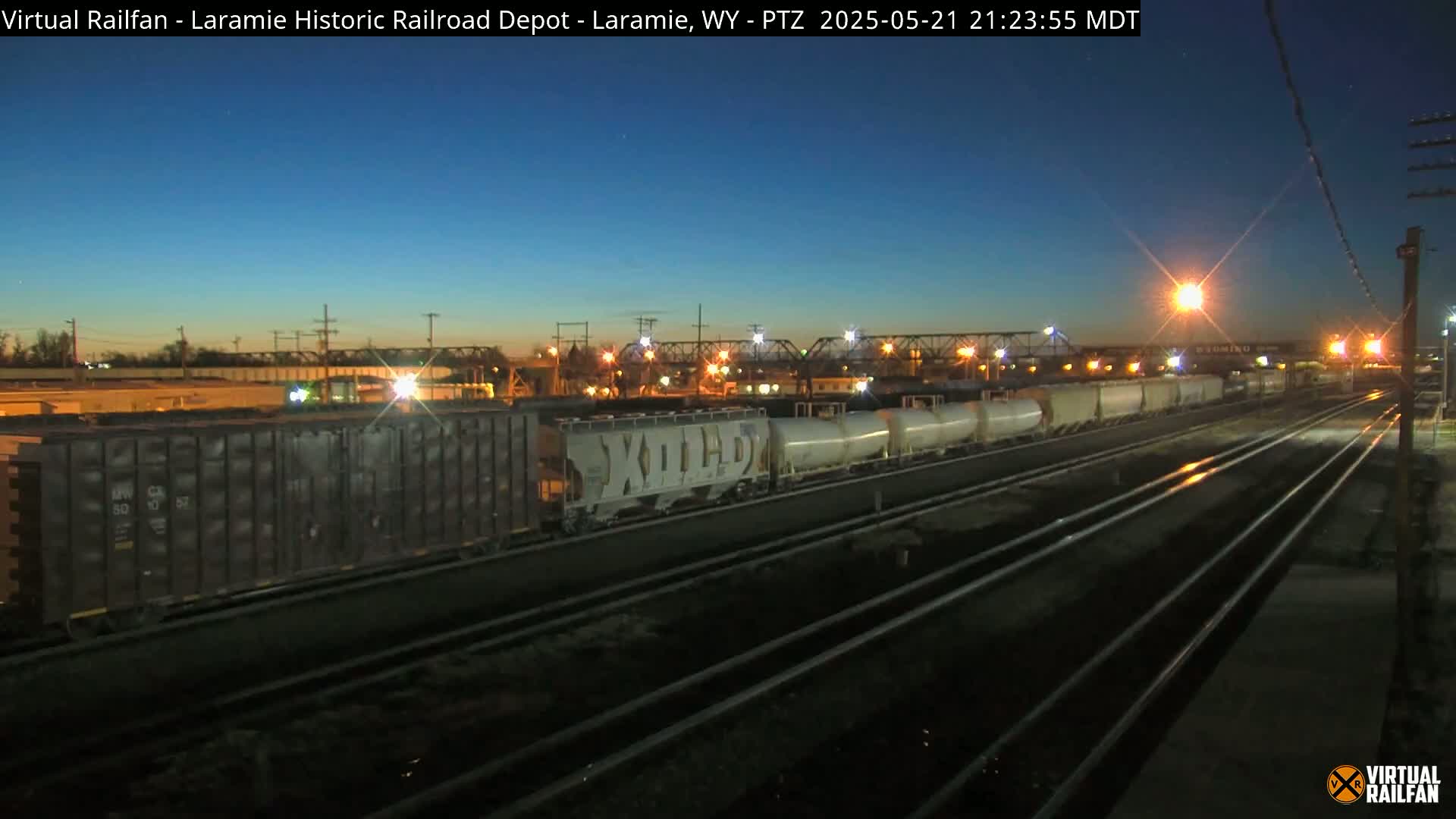 Under a twilight sky, a long freight train sits on railroad tracks, lit by nearby lights.