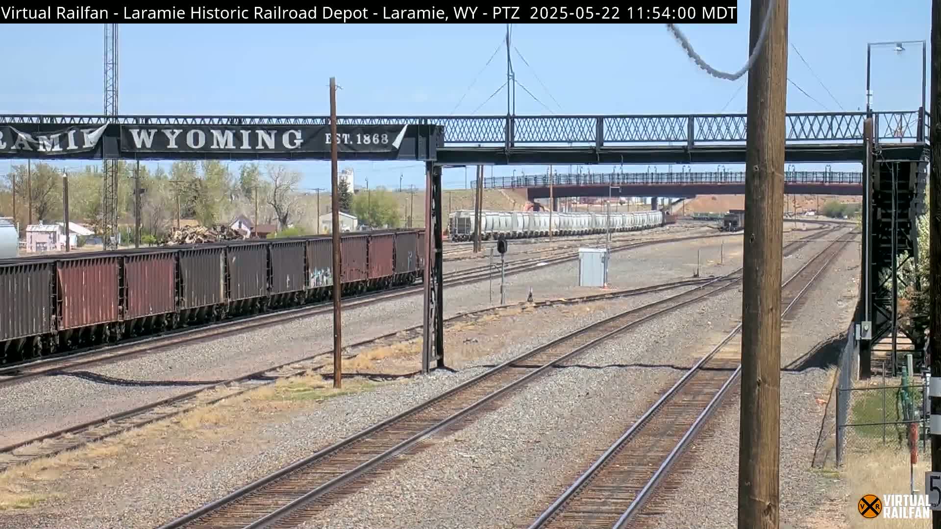 Under a clear sky, several freight trains are visible on a railway network that passes under a pedestrian bridge marked "Laramie, Wyoming."