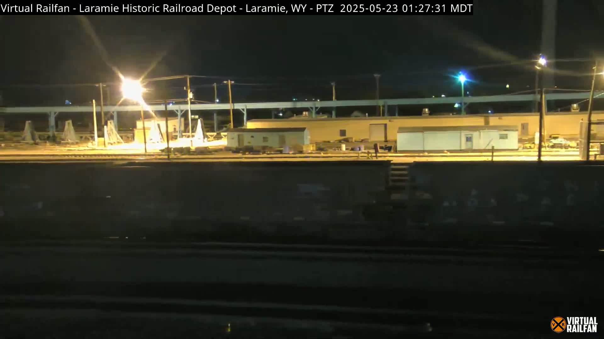 At night, a train car sits in front of a dimly lit rail yard with several buildings and structures.