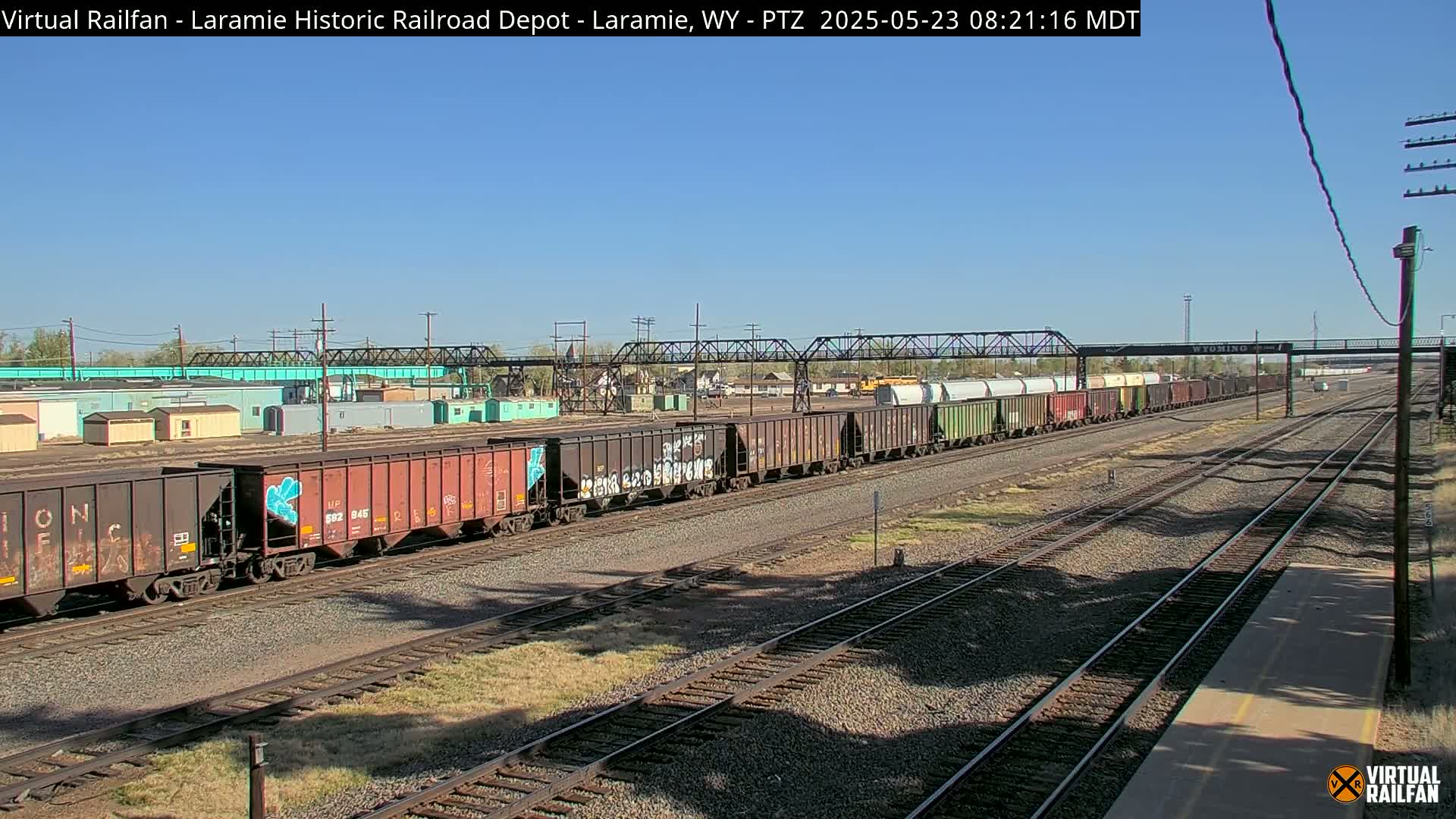 A long freight train, some cars of which are graffitied, sits on a rail yard under a clear blue sky.