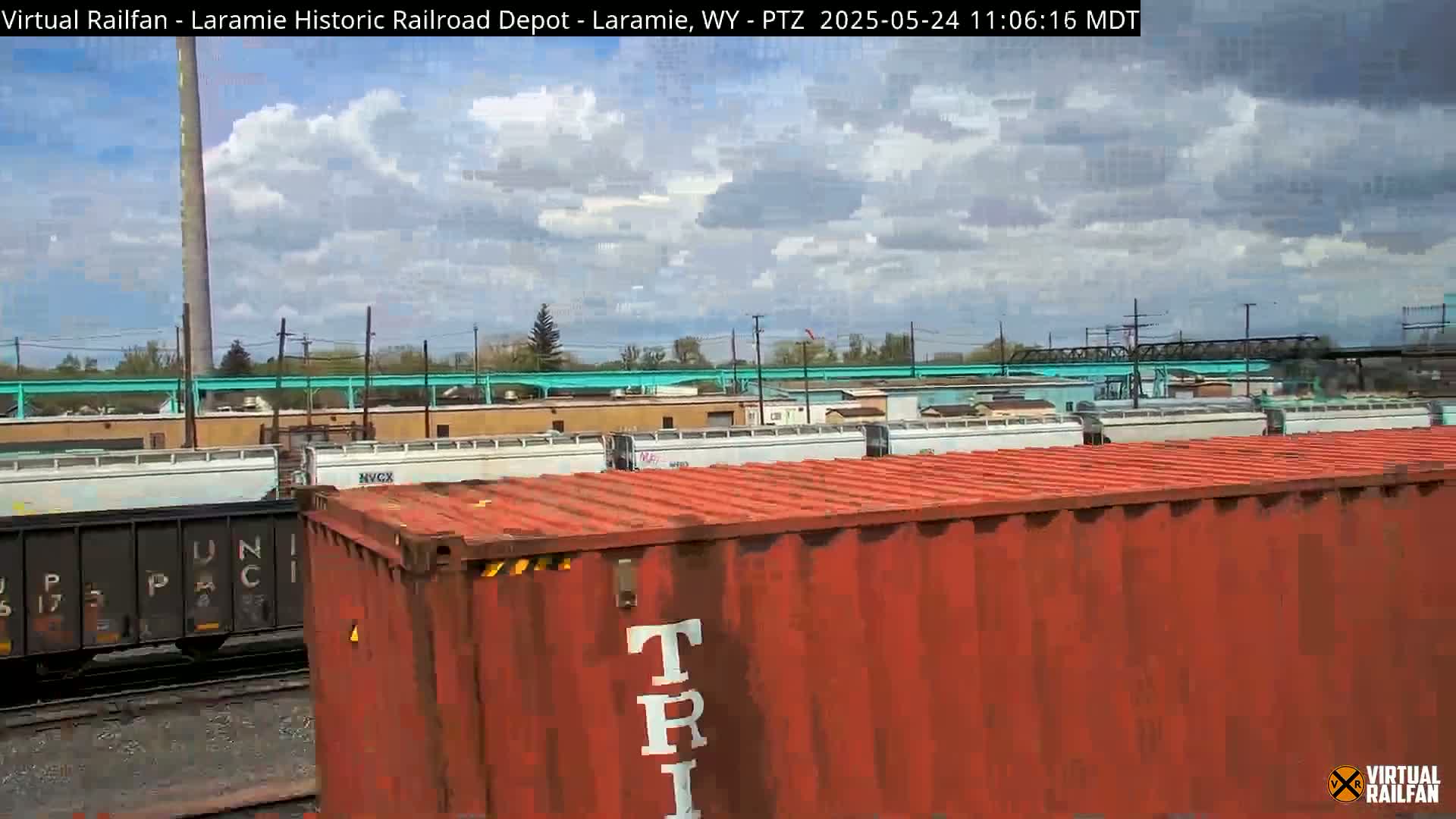 A partly cloudy day reveals a red shipping container in the foreground, with several railroad cars and industrial buildings visible in the background.