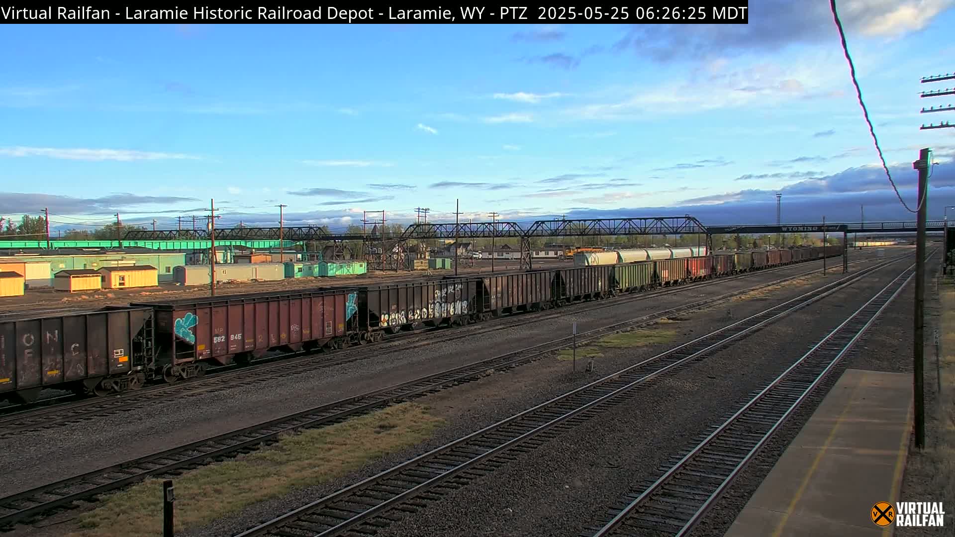 A long freight train sits on railroad tracks under a mostly clear, blue sky with a few clouds.